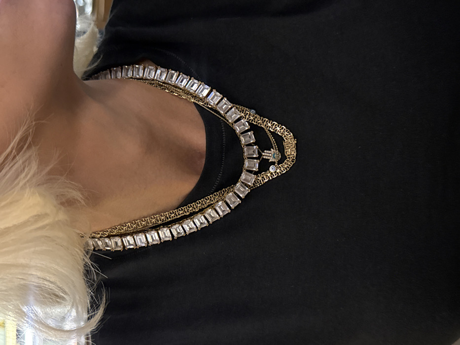 The image displays the upper torso and lower facial profile of an individual with short, platinum blonde hair and light skin, viewed from the right shoulder towards the mid-chest. The subject is wearing a plain black round-neck top. Adorning the neck are multiple layered gold-toned necklaces. The innermost necklace features a series of rectangular, clear, faceted stones set in a continuous band. Below this are at least two thinner gold-toned chains. One chain includes a small Hamsa hand charm with a central blue 'evil eye' motif. Another chain features a rectangular gold-toned pendant with an inscription that is not clearly decipherable but may relate to "DUBAI." The background is out of focus, indicating an indoor setting with indistinct features. The exact location within Dubai, United Arab Emirates, is not visually discernible from the scene itself.