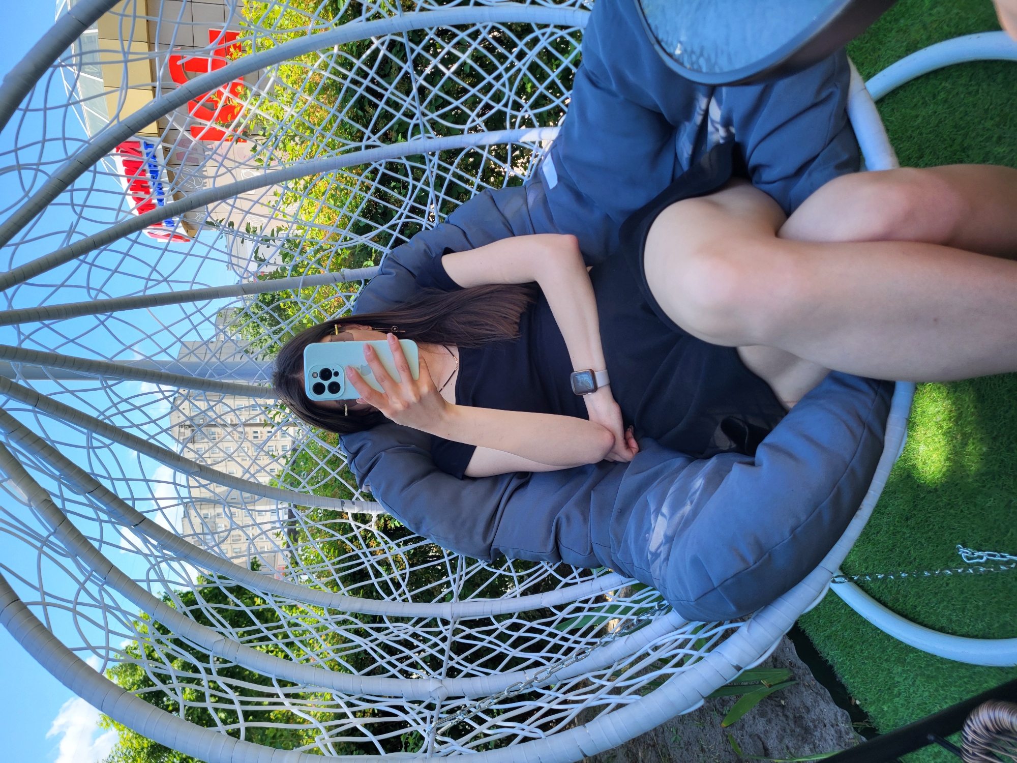 A young woman with long, dark hair is sitting in a large, white, egg-shaped swing chair, partially obscured by the phone she is holding up to take a selfie. She is wearing a black dress and a grey jacket, with a smartwatch on her left wrist. The chair is suspended by chains and features a large, plush grey cushion. Behind her, a building with a large red and white sign is visible, along with greenery and a blue sky with scattered clouds. The ground beneath the chair is covered in artificial green turf. The setting appears to be an outdoor seating area in Horishni Plavni, Ukraine, likely at a cafe or public space.