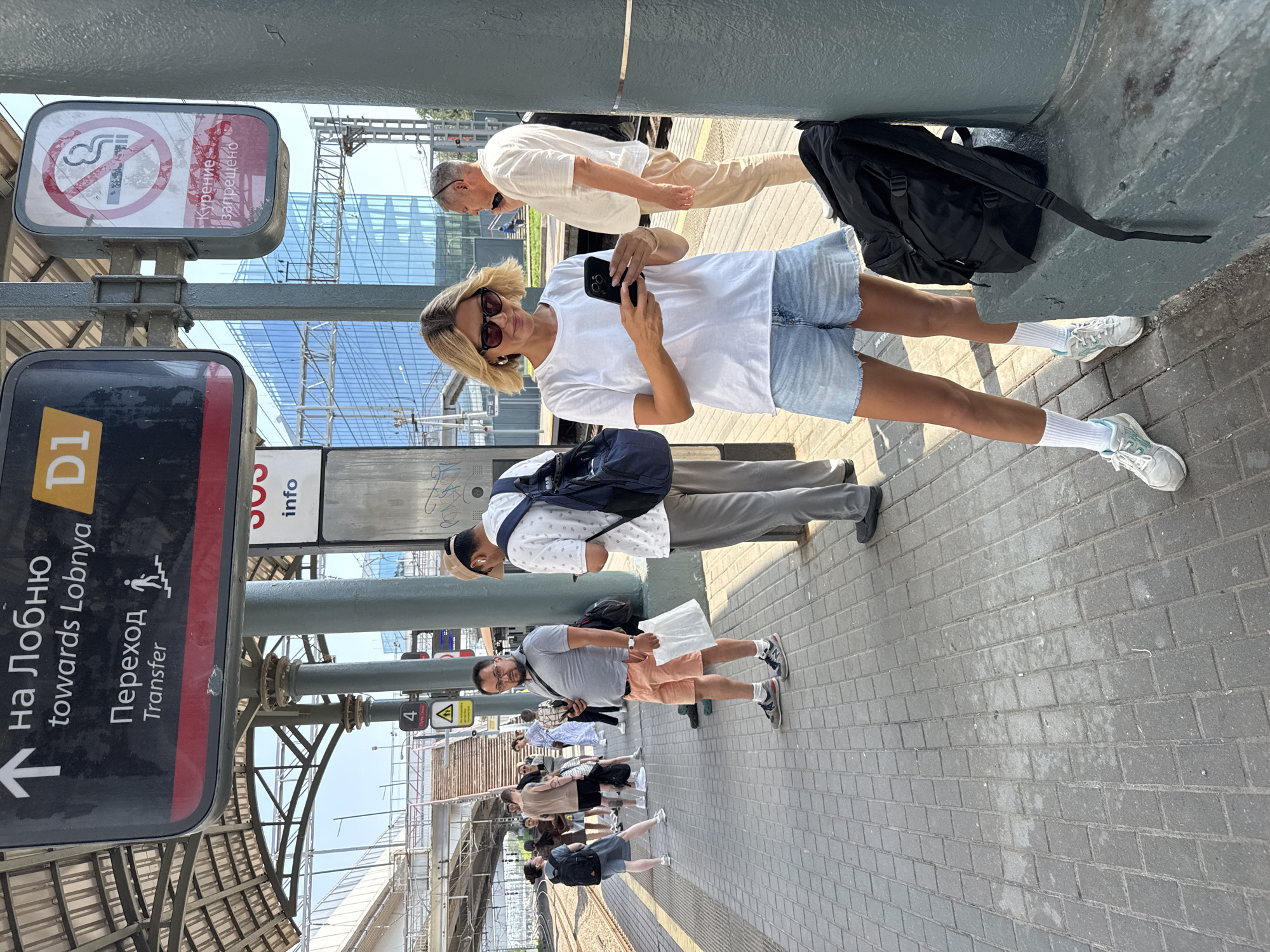 The image depicts a train platform in Khimki, Russia, under a covered structure. In the foreground, a blonde woman with sunglasses is looking at her smartphone. She is wearing a white t-shirt, denim shorts, white socks, and athletic shoes. A black backpack is leaning against a pillar next to her. To her right, a man in a white short-sleeved shirt and beige pants is talking on his phone. In the mid-ground, several people are waiting on the platform, including a man in a grey t-shirt and orange shorts holding a white bag, and a man in a grey shirt and black backpack. A sign overhead indicates "Towards Lobnya" and "Transfer." A "No Smoking" sign is also visible. The platform is paved with interlocking bricks, and in the background, a modern building with a glass facade and a train track can be seen.
