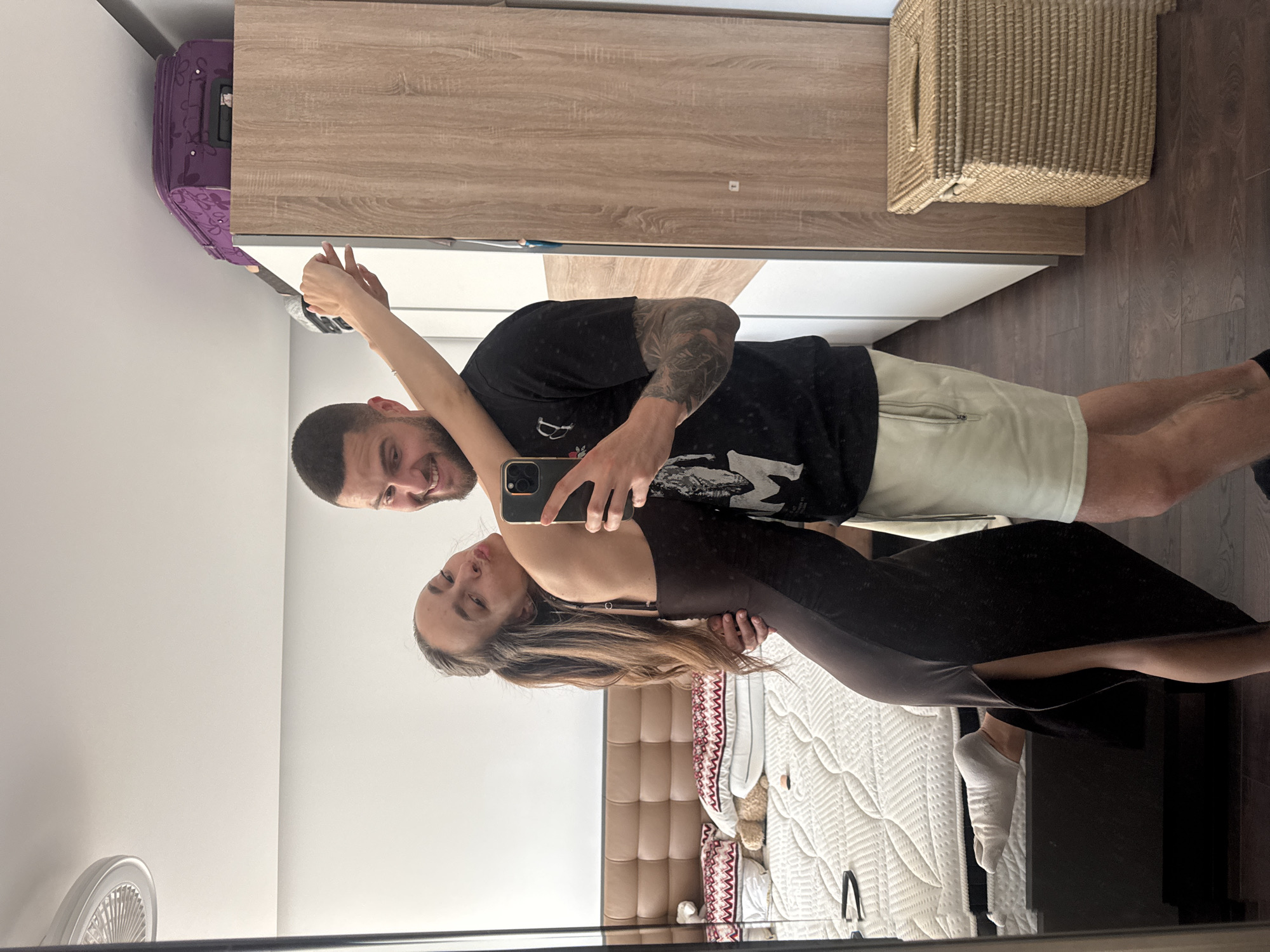 A selfie is taken in front of a bedroom mirror. A man with short dark hair, a beard, and tattoos on his arm holds a smartphone. He is wearing a black t-shirt and light-colored shorts. Beside him is a woman with long brown hair, wearing a dark dress. The bedroom includes a bed with a white and beige quilted bedspread, a beige headboard, a closet with light wood doors, a purple suitcase on top of the closet, a wicker storage box, and dark wood floors. A white wall is visible, and a fan is mounted on it. There is no specific information in the image that indicates the location is in Sofia, Bulgaria.