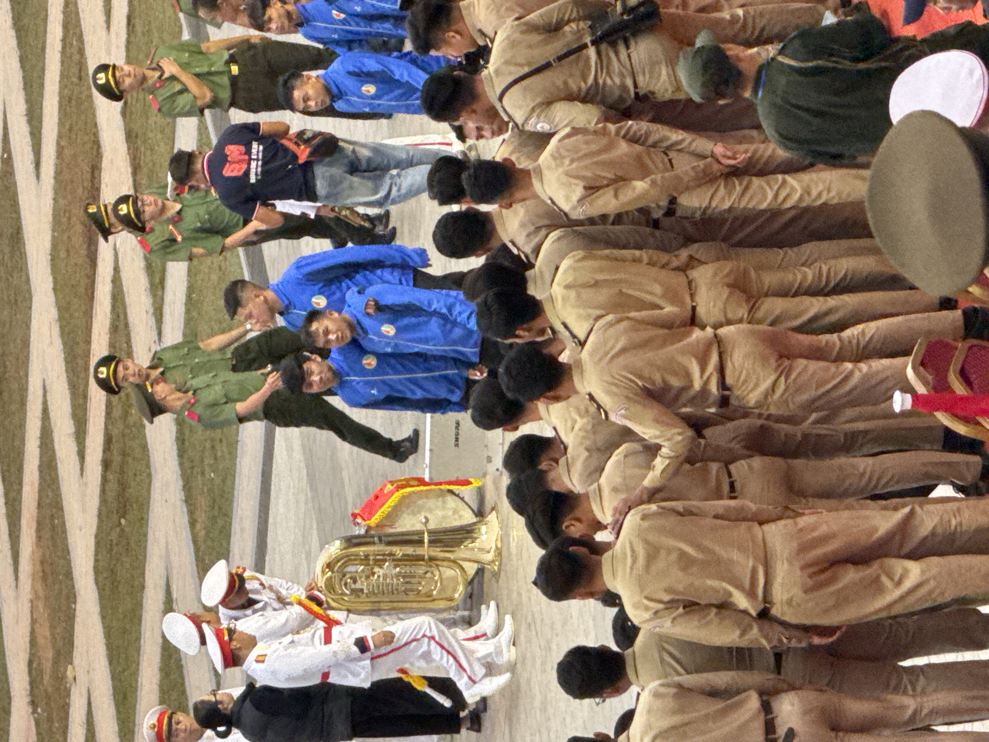 This is a photograph taken in Da Lat, Vietnam, depicting a marching band or ceremonial group. The subjects are primarily young men dressed in military-style uniforms. The uniforms vary, with some being green and others a tan or khaki color. Many of the individuals are wearing hats, some of which are black with gold insignia, and others are white with red trim.
The group appears to be in formation, with many individuals standing at attention or in a parade-like posture. Several people are holding musical instruments, most notably a large brass tuba prominently placed in the foreground. Others are holding batons or have their arms positioned in a formal manner. The setting appears to be an outdoor, paved area, possibly a plaza or street, with light-colored lines marking the ground.
The photograph is taken from a slightly elevated angle, looking down on the group. The lighting suggests it is daytime, though the weather is not clearly discernible. The expressions on the faces that are visible are neutral or serious, consistent with a formal occasion.
There is some visible text on what appears to be a piece of equipment in the background, partially obscured, with the letters "SHURE" visible. The overall scene conveys a sense of order and discipline, likely related to a public event or ceremony in Da Lat.