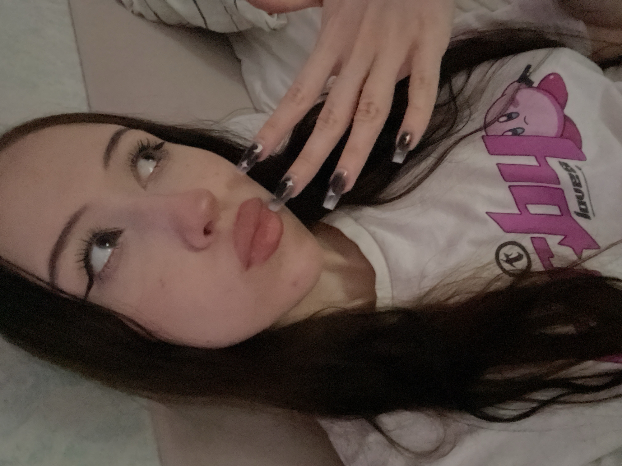 The image is a close-up, self-taken photograph of a young woman with long, dark hair, wearing a white t-shirt with a pink "Kirby" graphic. She has dramatic winged eyeliner and her lips are slightly parted, with her tongue visible. Her left hand, with long, dark-tipped, clear acrylic nails featuring small star designs, is raised near her face. The background appears to be a soft, neutral-colored surface, possibly a bed or sofa, with a striped pillow visible on the right. There is no indication of the location in Votkinsk, Russia, or any specific environmental details beyond the immediate indoor setting.