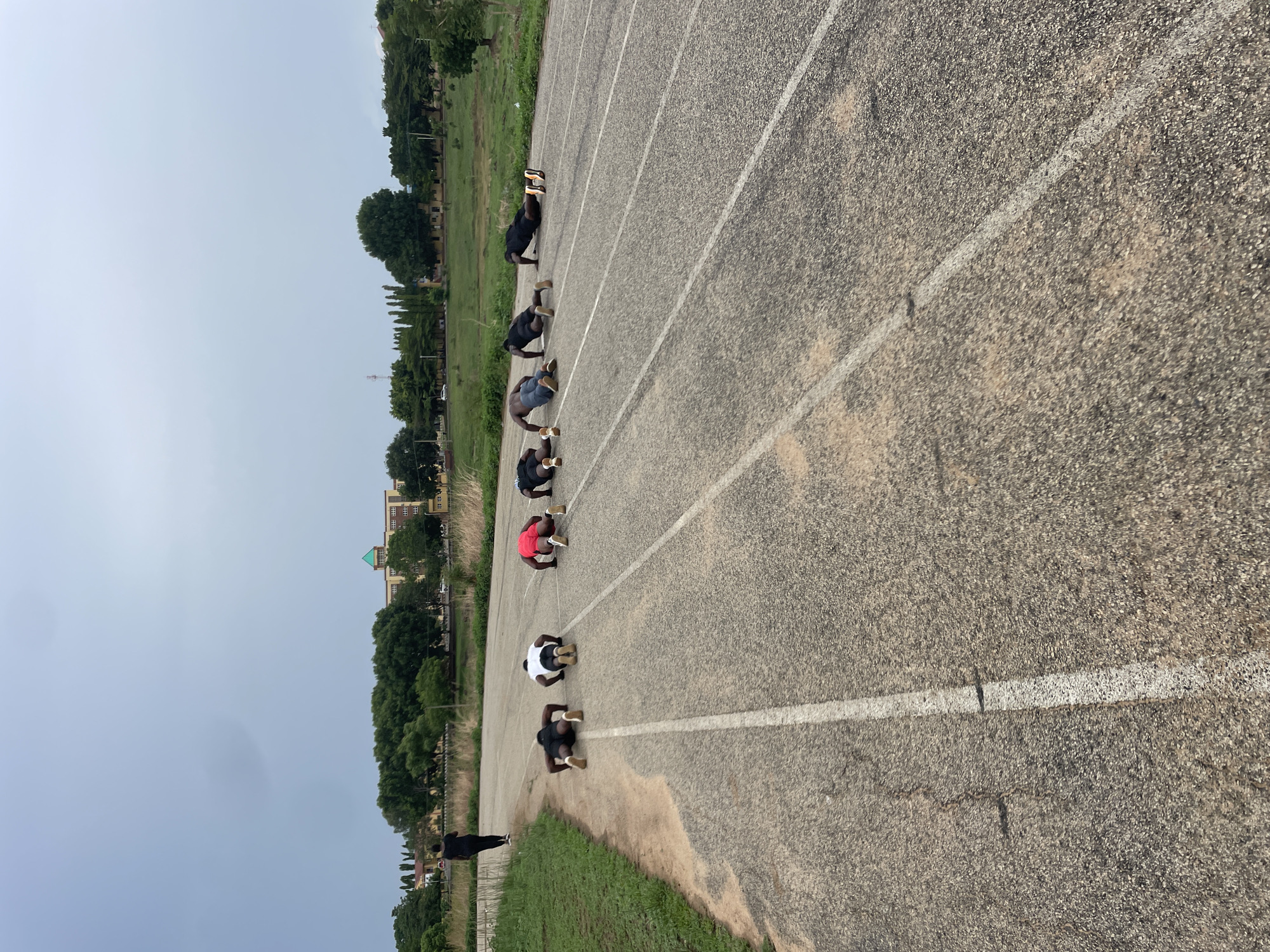 A photo captures a ground-level view of seven individuals positioned along a track field, performing push-ups in a straight line, each within their respective lane. To the left, a slender figure dressed in black stands observing the group. The track is marked with faded white lines, and the texture of the asphalt shows wear. Adjacent to the track, grass borders the edge. Beyond the grassy area, trees are visible, lining the horizon. In the distance, a building with a green-tiled roof is present. The sky overhead appears overcast. The photograph was taken in Keffi, Nigeria.
