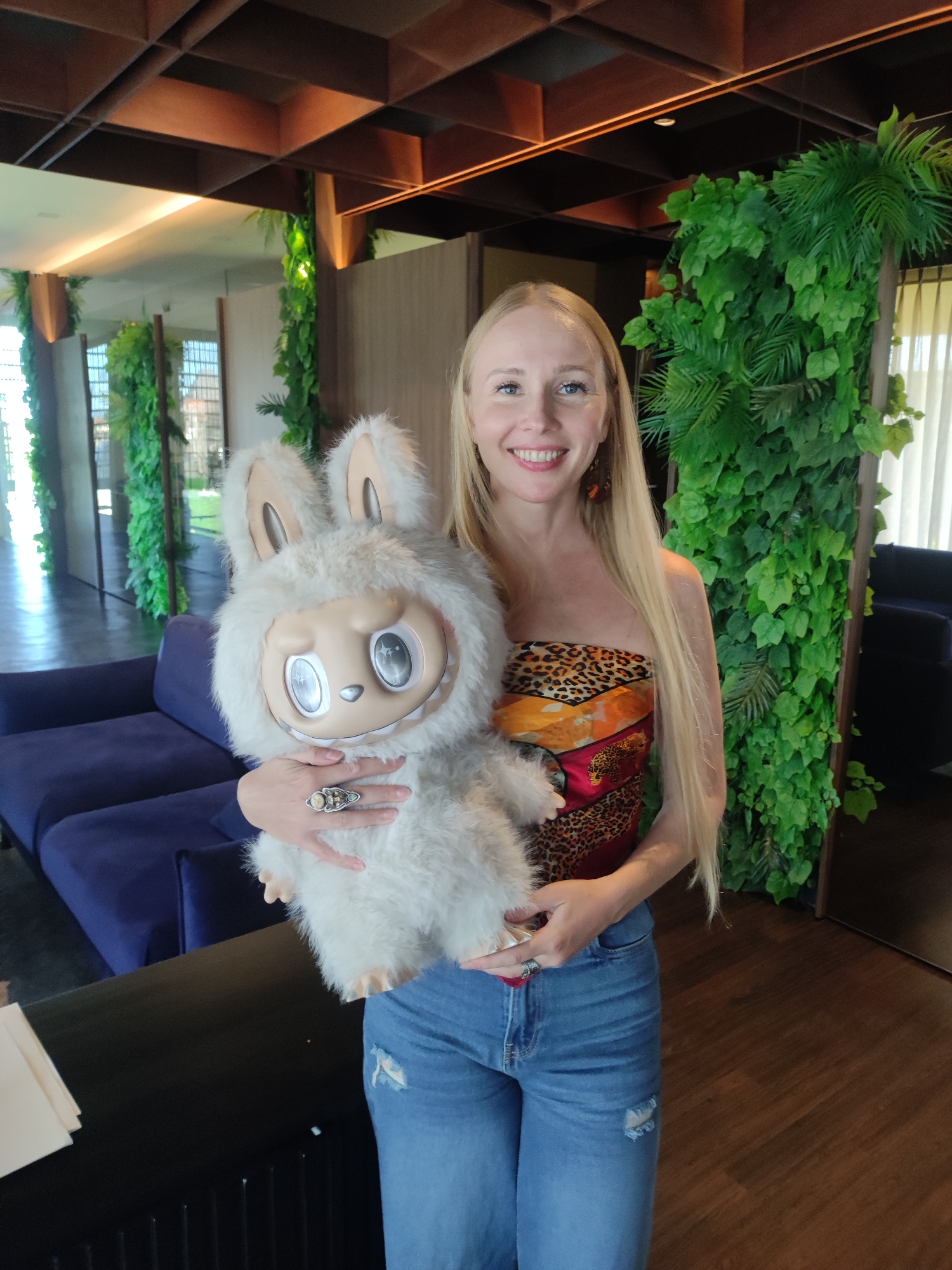 A fair-skinned woman with long blonde hair stands indoors, holding a furry white rabbit-like doll. She is wearing a strapless top with a leopard print and red accents, and light blue jeans with a ripped knee. She has a large ring on her right hand. The doll has large, round, shiny eyes and a neutral expression.
The background includes a blue sofa, a dark table or countertop, and walls decorated with green foliage. There are wooden beams on the ceiling. The location is North Kuta, Indonesia.