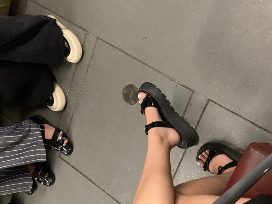 The image presents a ground-level view of the lower extremities of at least three individuals on a grey tiled floor. In the foreground, a person is seated, with their bare legs visible above a red, padded surface, and their feet clad in black patent leather platform sandals featuring thick, ridged soles and bow embellishments on the straps. The toenails on the visible foot are painted light blue. To the upper-left, another individual stands, wearing vertically striped trousers and black, multi-strapped sandals with closed toes. To the upper-right, a third person stands, dressed in wide-leg black trousers and cream-colored flat shoes with substantial, rounded soles. The floor is composed of large, rectangular grey tiles with visible grout lines. A small, circular, dark metal object, possibly a utility cover, is embedded in the tile surface towards the center-right of the frame. No specific architectural or environmental details are present to confirm a precise location within the city of Kozyn, Ukraine.