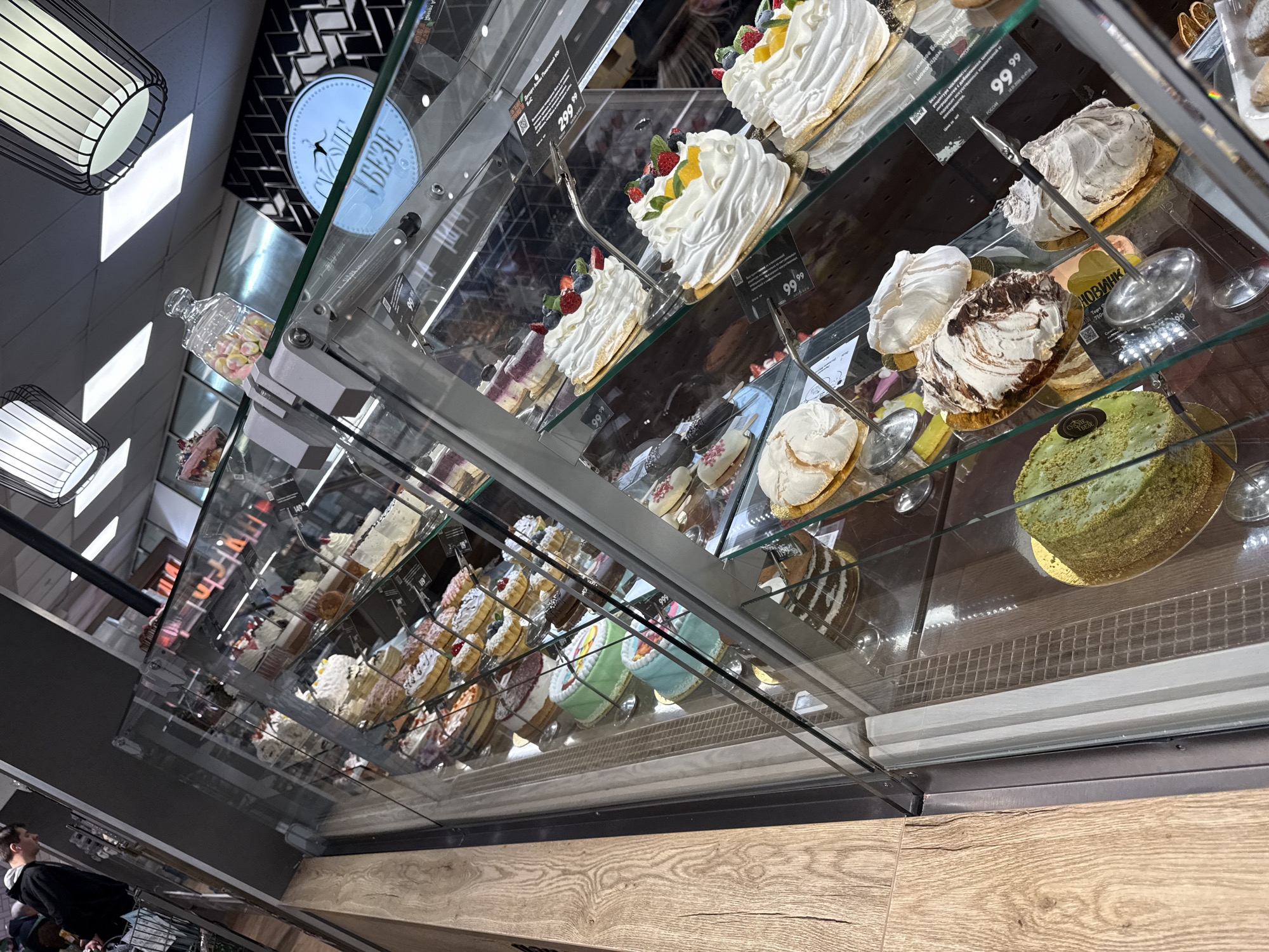 A bakery display case is filled with various cakes and pastries. Many of the cakes are topped with white frosting and fresh fruit. Price tags are visible on the shelves. The display case is located in Krasnoyarsk, Russia, as indicated by the Russian text on some of the price tags. A person is partially visible in the lower left corner of the image. The background includes lighting fixtures and wall decorations. The counter in front of the display case is made of wood.