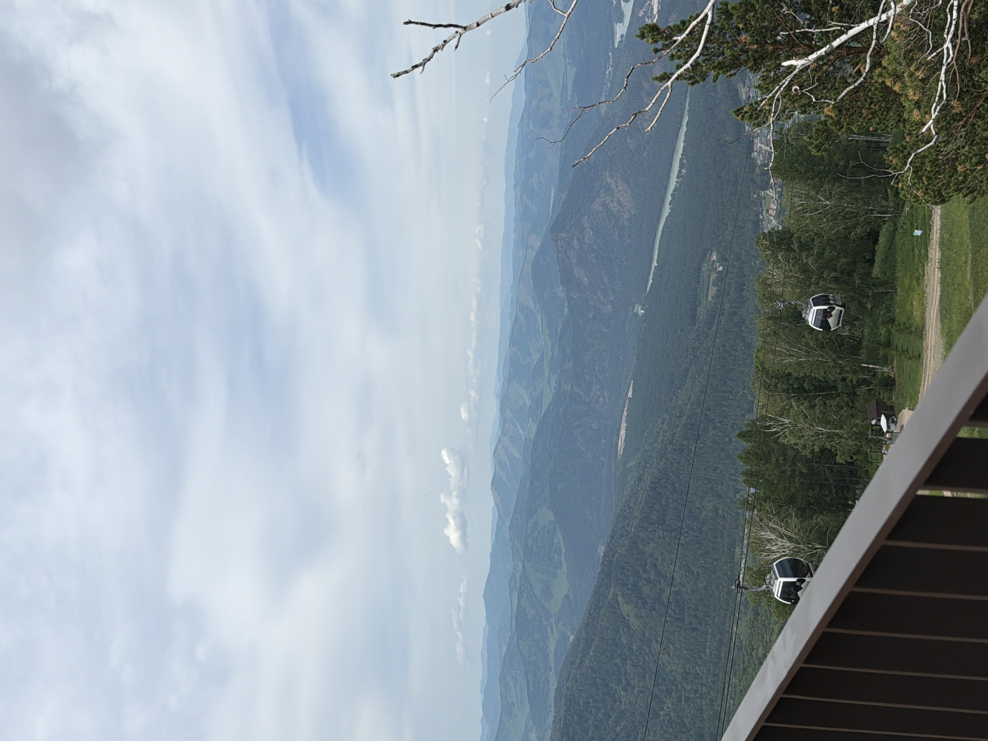 The image captures a scenic view of rolling green mountains under a cloudy sky. In the foreground, a dark railing partially obstructs the view of a ski lift or cable car ascending a forested slope. A white cable car is visible suspended on the cable. The middle ground features dense green forests covering the mountainside, with a river winding through the valley. In the distance, layers of blue-hued mountains stretch towards the horizon. The location is Ozernoe, Russia.