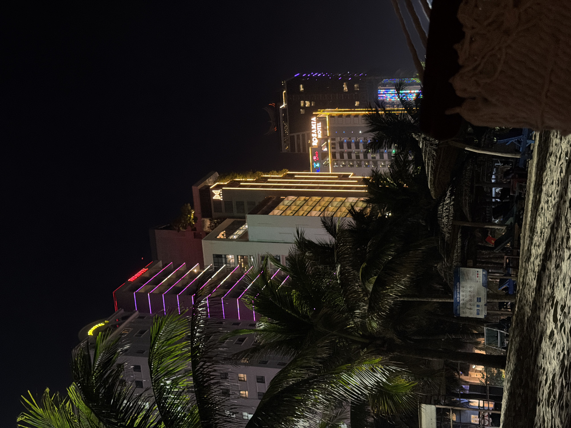This nighttime photograph captures a vibrant cityscape in Da Nang, Vietnam, bustling with illuminated buildings and tropical foliage. The image, which appears to be rotated 90 degrees clockwise from its intended landscape orientation, showcases a lively urban scene viewed from an elevated perspective, possibly a balcony or a raised outdoor area.
In the foreground, a textured, light-colored surface, resembling sand or a stone wall, occupies the right side, with a piece of woven, light-colored fabric or rope hanging into the frame from the top right. Numerous palm trees, their fronds silhouetted and partially lit by ambient light, are scattered across the midground and foreground, adding a distinctly tropical atmosphere.
The background is dominated by several modern high-rise buildings, brightly illuminated against the dark night sky. One prominent structure features an orange neon sign clearly displaying "ROSAMIA HOTEL" with "Bar" visible below it. Another building to the left presents red neon lighting that spells out what appears to be "TURI HOTEL" or a similar name. Other buildings are adorned with geometric patterns of yellow strip lights and distinctive purple, diagonally arranged neon lights, contributing to the dynamic visual appeal.
Further down and to the right, obscured by palm trees, a blue informational sign with white Vietnamese text and icons is visible. Partially readable phrases on the sign include "HỘI QUY TRẢ LỜI."
The scene is set at night under clear weather conditions, emphasizing the artificial lights of the city. The presence of hotels and illuminated establishments suggests a bustling tourist or entertainment district, although specific human activity is not clearly visible. The overall impression is one of a vibrant, modern, and inviting tropical city at night.