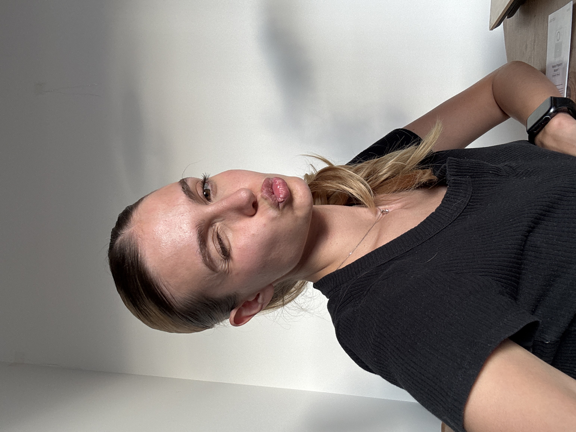 The image is a close-up portrait of a young woman posing playfully, captured in a selfie-style shot. She is seen with her head tilted, making a "kissing" or "duck face" expression with her lips pursed. One eye is partially closed or squinted, while the other is open, revealing a brown iris. Her blonde hair, with some darker roots, is pulled back.
She is wearing a black, ribbed short-sleeved top and a thin silver chain necklace. A black digital smartwatch is visible on her left wrist.
The background consists of a plain, light-colored wall, which shows some subtle shadows and faint smudges. In the upper right corner, a wooden surface, possibly a shelf or furniture, is visible, along with a white piece of paper. Faint text on this paper can be discerned, including words that resemble "Jumbo" and "Rinco Proces," as well as a numeric "0" icon. The lighting is bright and even, suggesting an indoor setting during the daytime. The overall mood is casual and expressive.