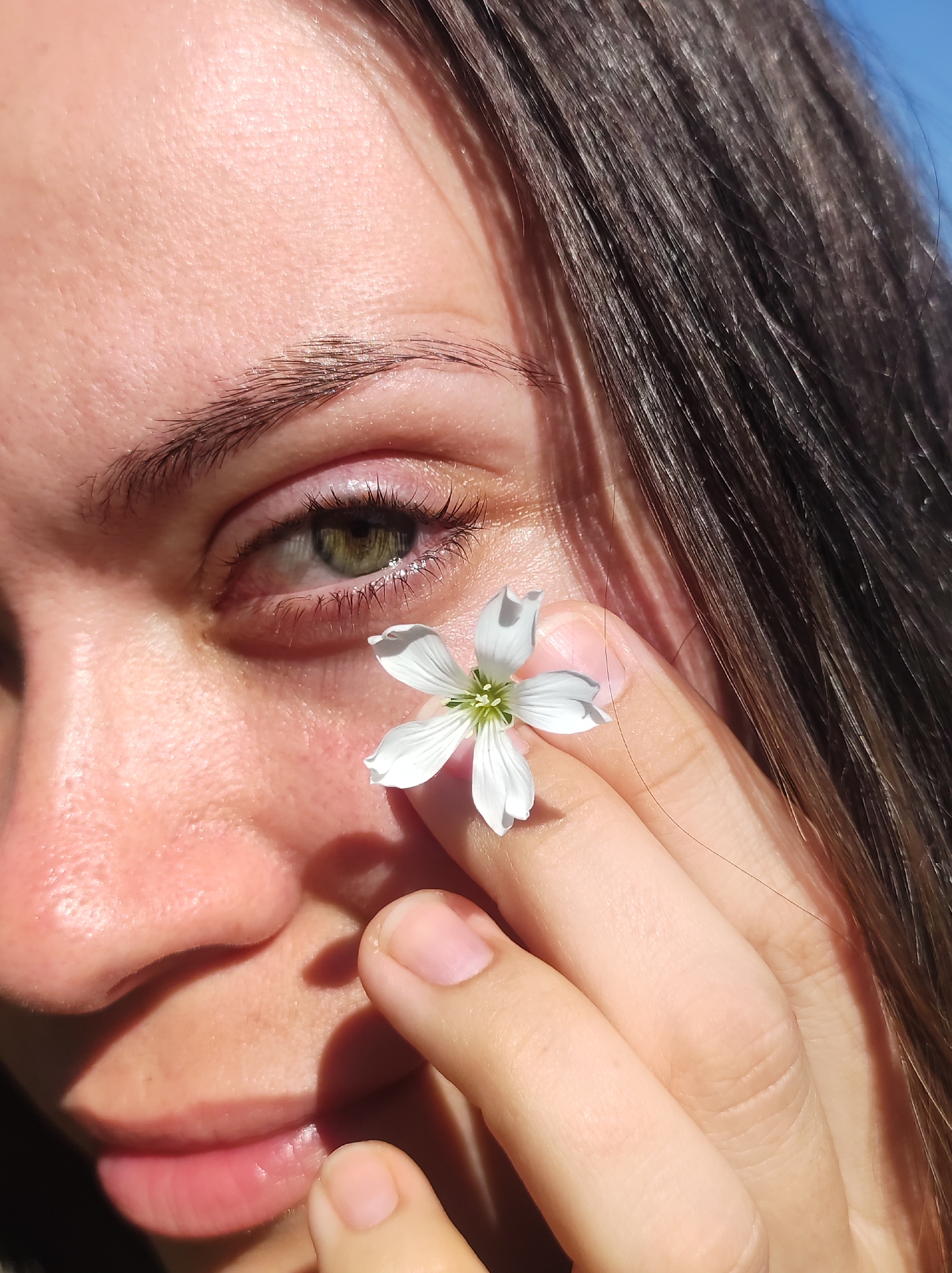 A close-up shot focuses on a person's face, specifically their left eye and cheek, as they hold a small white flower near their face with their right hand. The individual has dark hair, fair skin, and green eyes. The lighting suggests it is daytime, with shadows cast across the face, indicating sunlight. The background is blurred but appears to be an outdoor setting. There is no visible information to suggest the location is in Фоки, Russia.