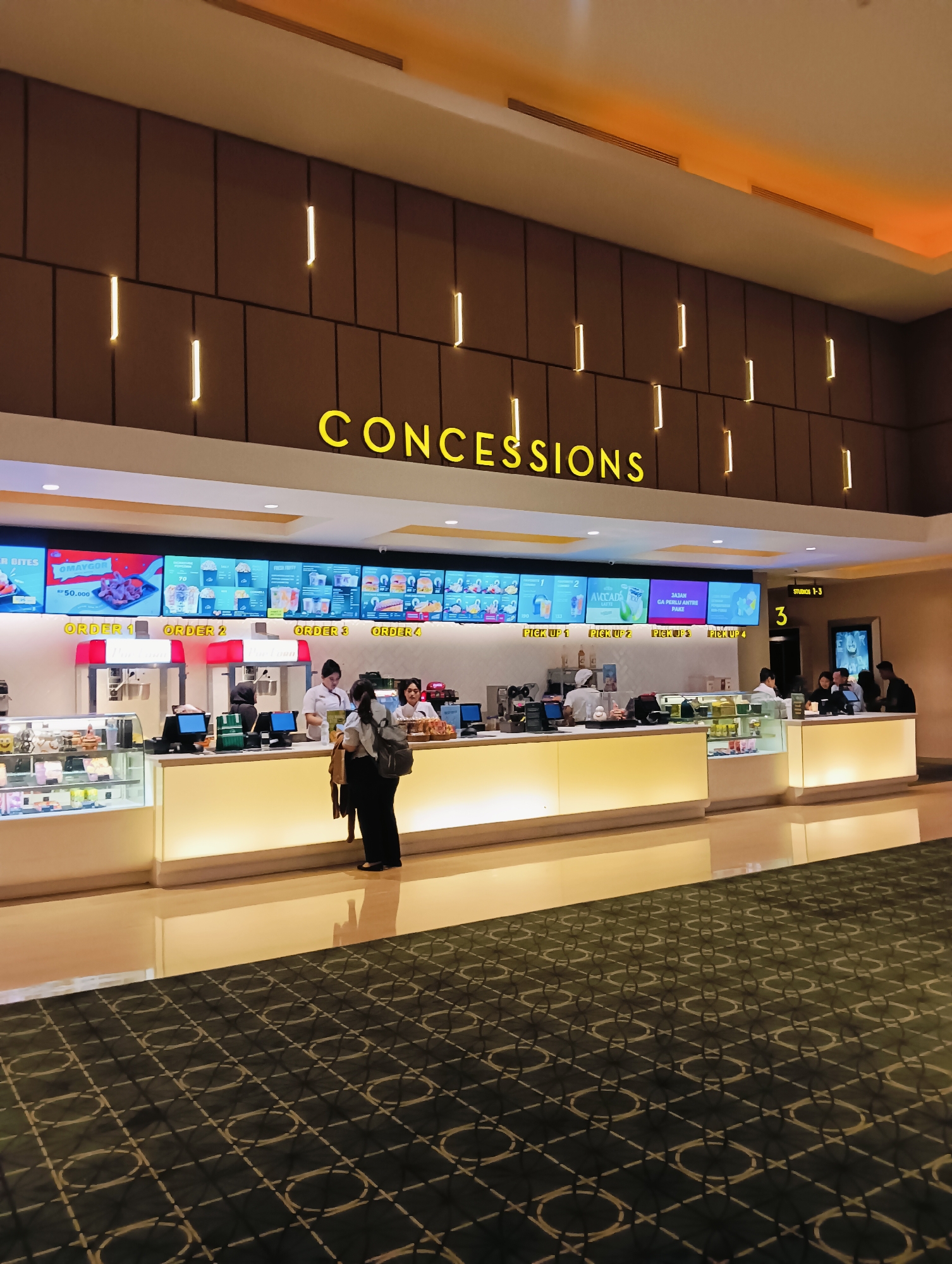 This image depicts the concessions area of a cinema, likely in Batam, Indonesia, given the presence of Indonesian language on the menu boards. The scene features a long, brightly lit counter where customers are ordering and picking up food and beverages. Several staff members in uniforms are visible behind the counter, serving customers. Digital menu boards display various food items and prices. In the foreground, a patterned carpet with an intricate design covers the floor. The overall atmosphere suggests a busy but organized concession stand within a modern cinema.