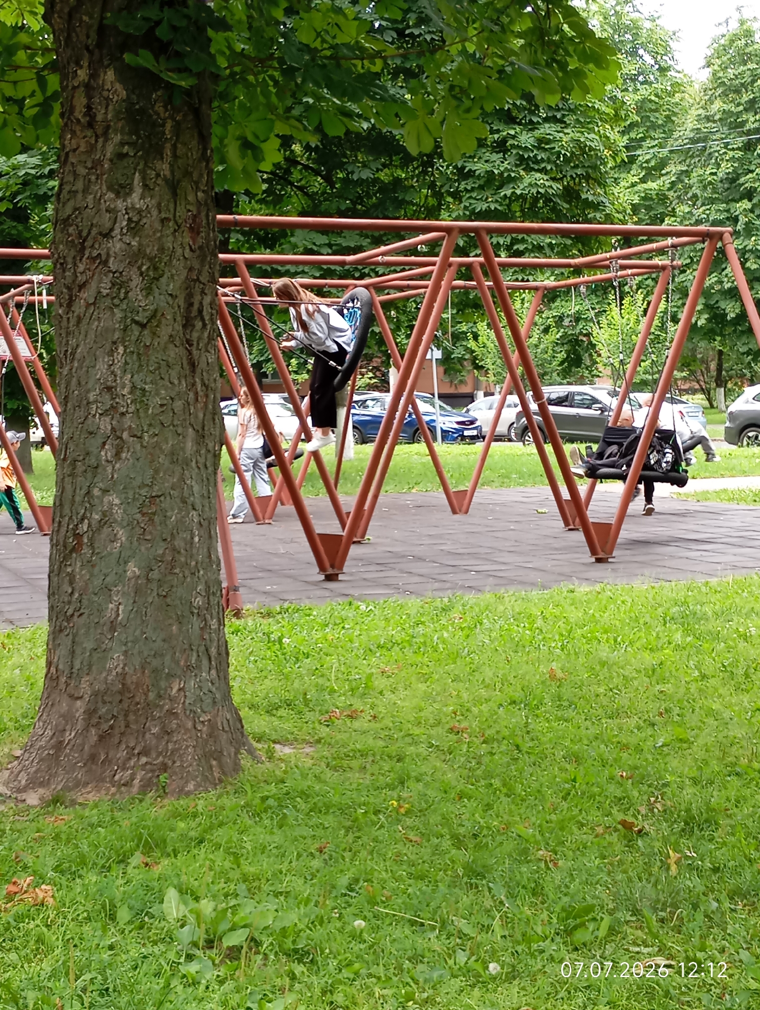 The image captures a playground scene in the city of Homyel, Belarus, featuring a swing set with multiple children on it. In the foreground, a large tree trunk is prominently visible on the left. The swing set, constructed from reddish-brown metal, is the central element of the playground. A young girl with long blonde hair, wearing a white and black top and black pants, is actively swinging. To her right, another child, with dark hair and wearing dark clothing, is also on a swing, leaning back. Further to the right, another person, possibly a child or teenager with dark clothing, is partially visible on a swing, with their legs extended. In the background, several cars are parked, suggesting the playground is located near a street or parking area. The ground beneath the swings is covered with dark rubber matting, and the surrounding area is covered in green grass. The overall environment appears to be an outdoor urban park or recreational area. The timestamp "07.07.2026 12:12" is visible in the lower right corner of the image.