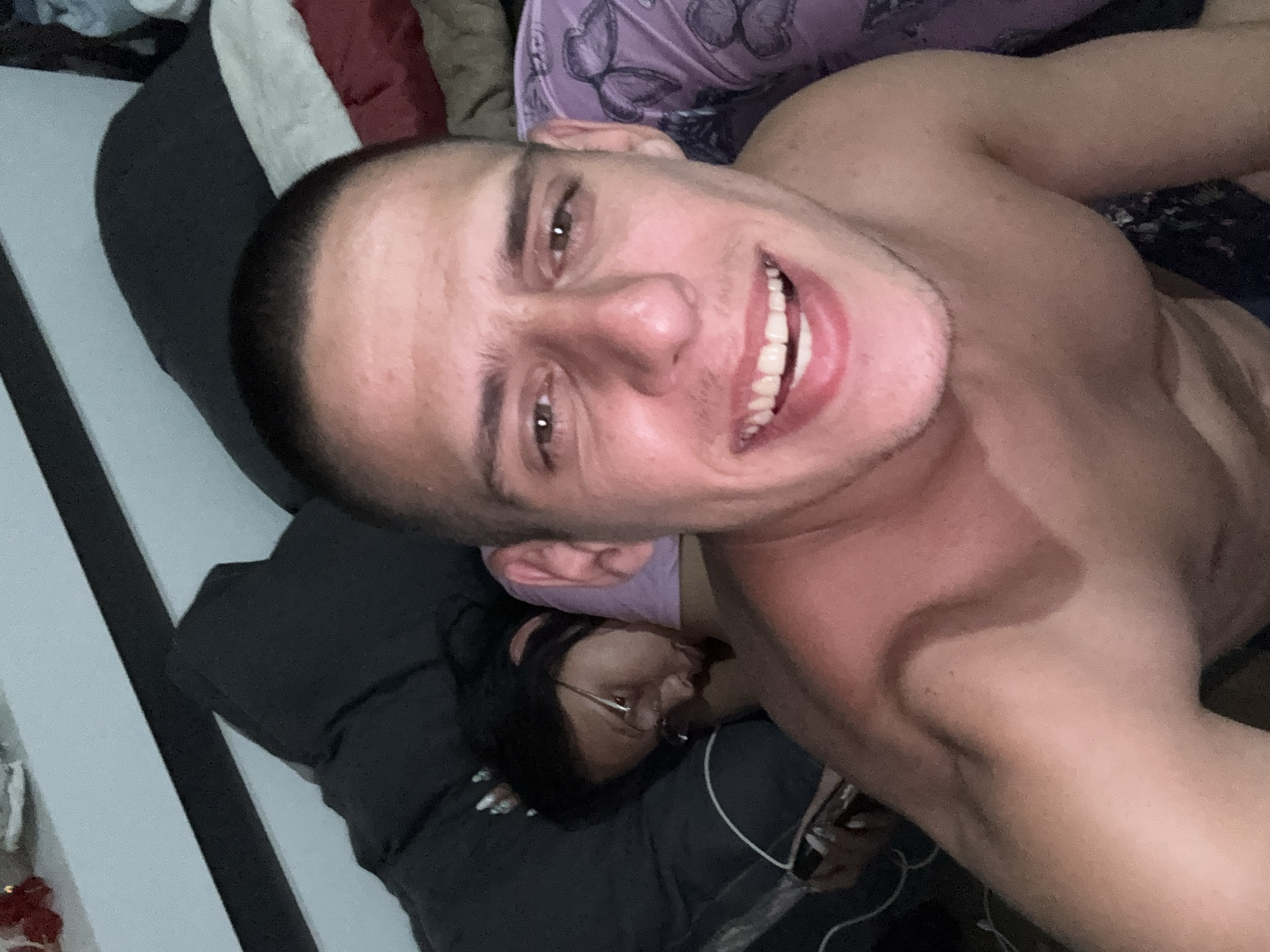 A shirtless man is lying on a bed, smiling broadly at the camera. Behind him, a woman with glasses is asleep on the bed. The location is unspecified, and there is no indication the image was taken in Sofia, Bulgaria.