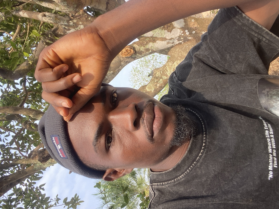 The scene depicts a close-up outdoor portrait of a young adult male, situated in the city of Igbarra, Nigeria. The subject has dark skin, short dark hair, dark eyebrows, and a dark goatee beard. He is wearing a dark grey beanie with a rectangular label displaying "abercrombie CALIFORNIA QUALITY GOODS" and a dark grey, possibly acid-washed, t-shirt. The t-shirt bears white printed text reading "No Matter Where Life Takes Me, Find Me With A Smile." His left hand is raised, fingers slightly curled, partially obscuring his forehead and the upper portion of his right eye. The subject's gaze is directed forward towards the camera. The environment consists of a natural background featuring tree branches with sparse green foliage and a bright, clear sky, indicative of daylight conditions.