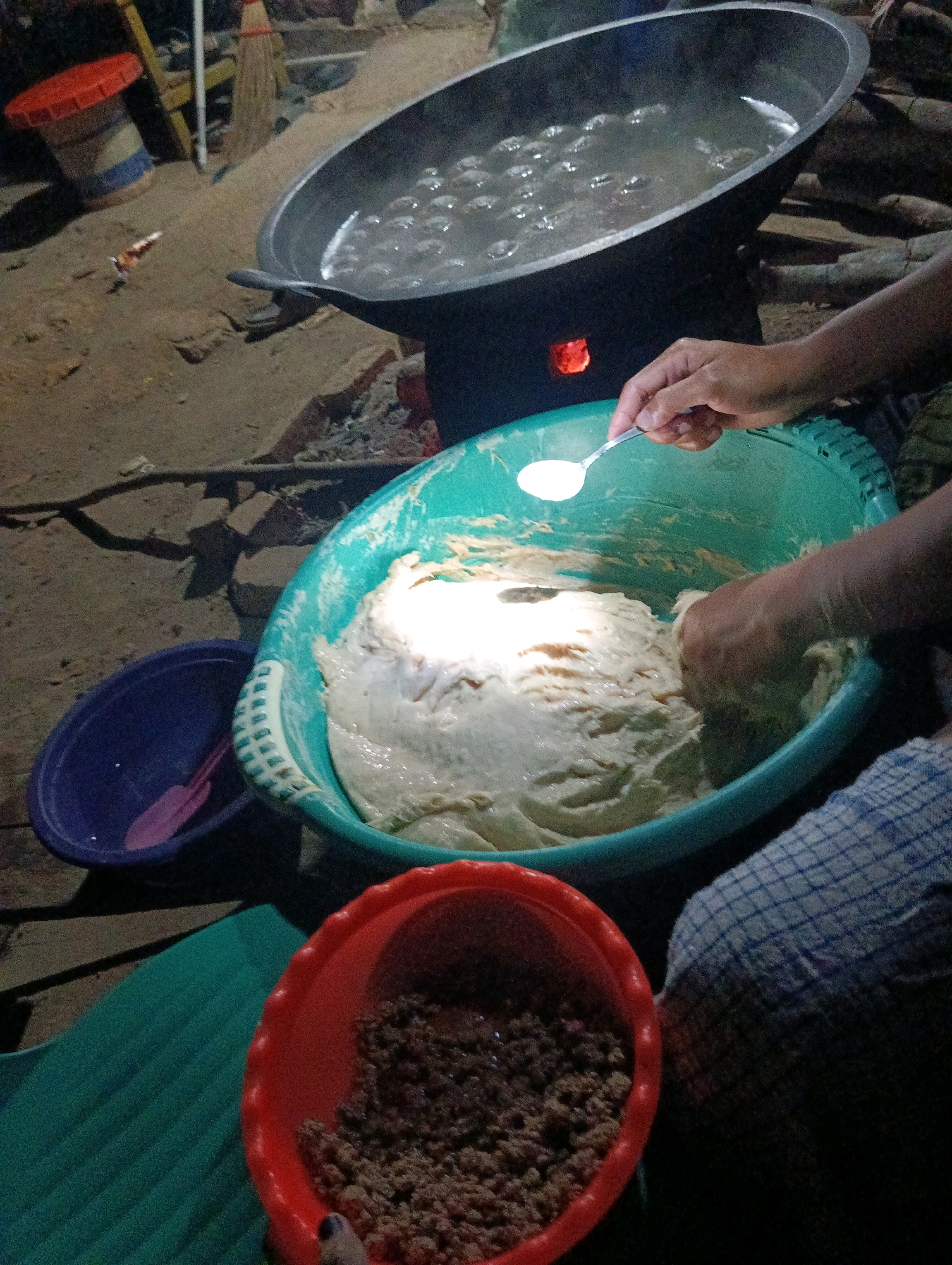 In Pematang Jaya, Indonesia, a scene of food preparation unfolds outdoors at dusk. A large, dark wok filled with boiling water and small, round objects is positioned over a fire fueled by burning wood. Beside it, a person's hands are visible, one holding a spoonful of white powder over a large teal bowl containing a light-colored, dough-like substance. Another hand is immersed in the dough, actively mixing it. To the left of the teal bowl is a smaller, dark blue bowl with a pink utensil inside. In the foreground, a bright red bowl holds a granular brown substance. The ground is dirt, with scattered debris, and a partial view of a red-lidded container and a broom can be seen in the background, suggesting a humble, outdoor cooking environment. A checkered blue and white cloth rests beside the teal bowl.