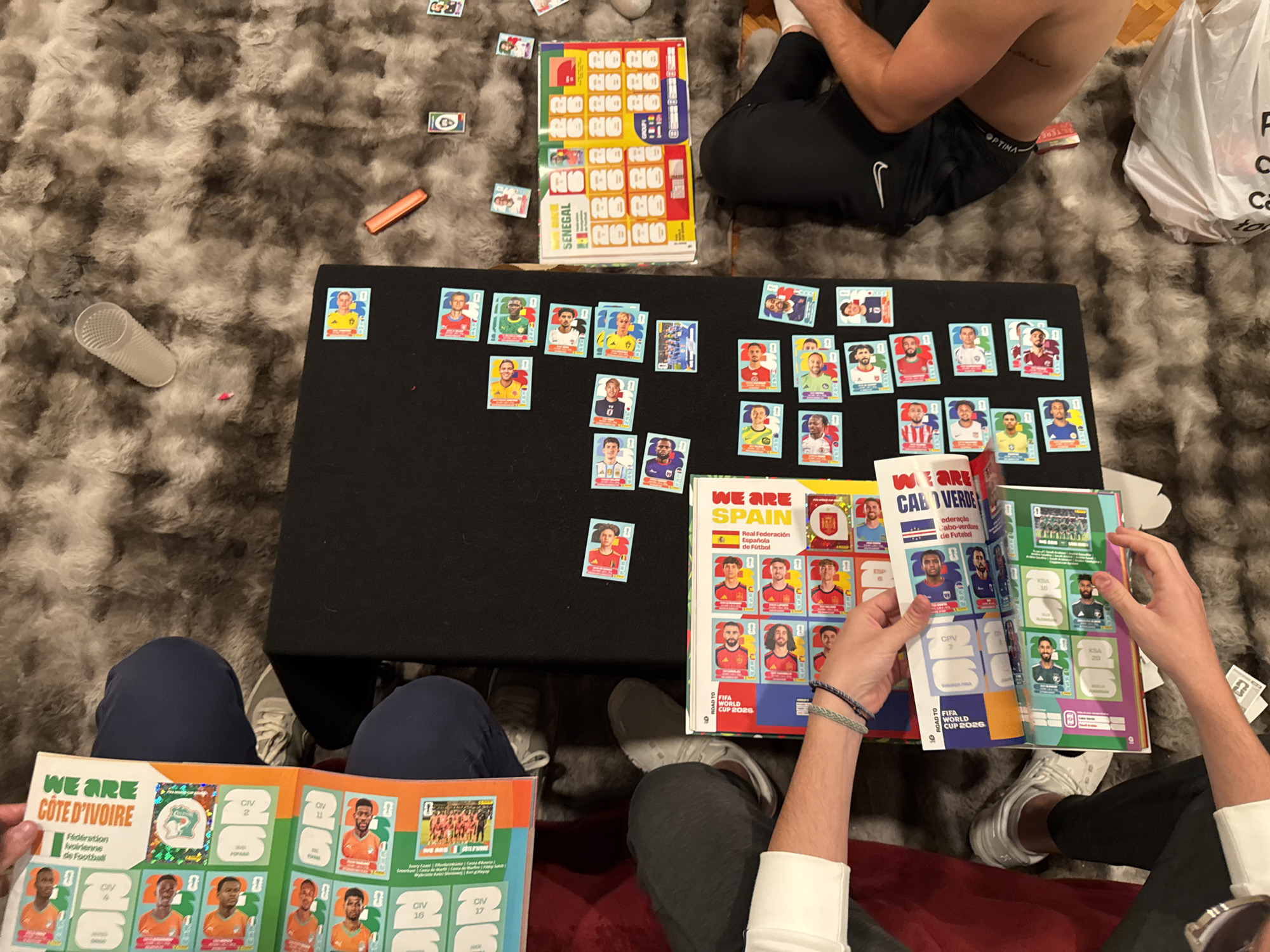The image shows three people sitting on a shaggy rug, engaged in a sticker album activity. Two of them are holding open different sticker albums titled "Côte d'Ivoire" and "Cabo Verde," while the third album lying on the table is of "Spain". A black table in the center holds a collection of loose stickers and sticker sheets labeled "Senegal". The person on the right is placing stickers into the “Cabo Verde” sticker album. A plastic cup and various small objects are scattered on the rug around the table. One person is shirtless with black pants. The location appears to be inside a home. There is nothing in the image to suggest the location is in Mexico City, Mexico.