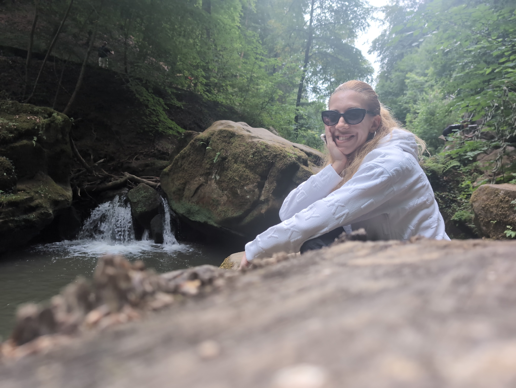 A blonde woman wearing sunglasses and a white hooded sweatshirt smiles at the camera, resting her chin on her hand. She is positioned on the right side of the frame, with a small waterfall and a body of water in the background. The environment is lush and green, with trees and moss-covered rocks surrounding the waterfall, suggesting a natural setting like a park or forest. A large, blurred log or rock is in the foreground, obscuring the bottom portion of the image. The scene appears to be outdoors and possibly in a humid environment, given the mistiness of the background.