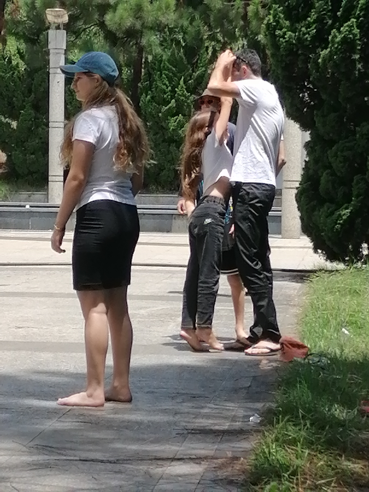 In Da Lat, Vietnam, four people stand on a paved pathway in a park. On the left, a young woman wearing a blue baseball cap, a white t-shirt, and a black skirt stands barefoot, facing left. Her long brown hair is visible extending past her shoulders. To her right, a younger girl in a white t-shirt and black jeans stands barefoot, facing forward. Next to her, a man in a white t-shirt and black pants also stands barefoot. A fourth person, partially obscured, stands behind the man and the younger girl. In the background, lush green trees and foliage surround the pathway, and a white pillar is visible to the left. The ground is a textured grey pavement.
