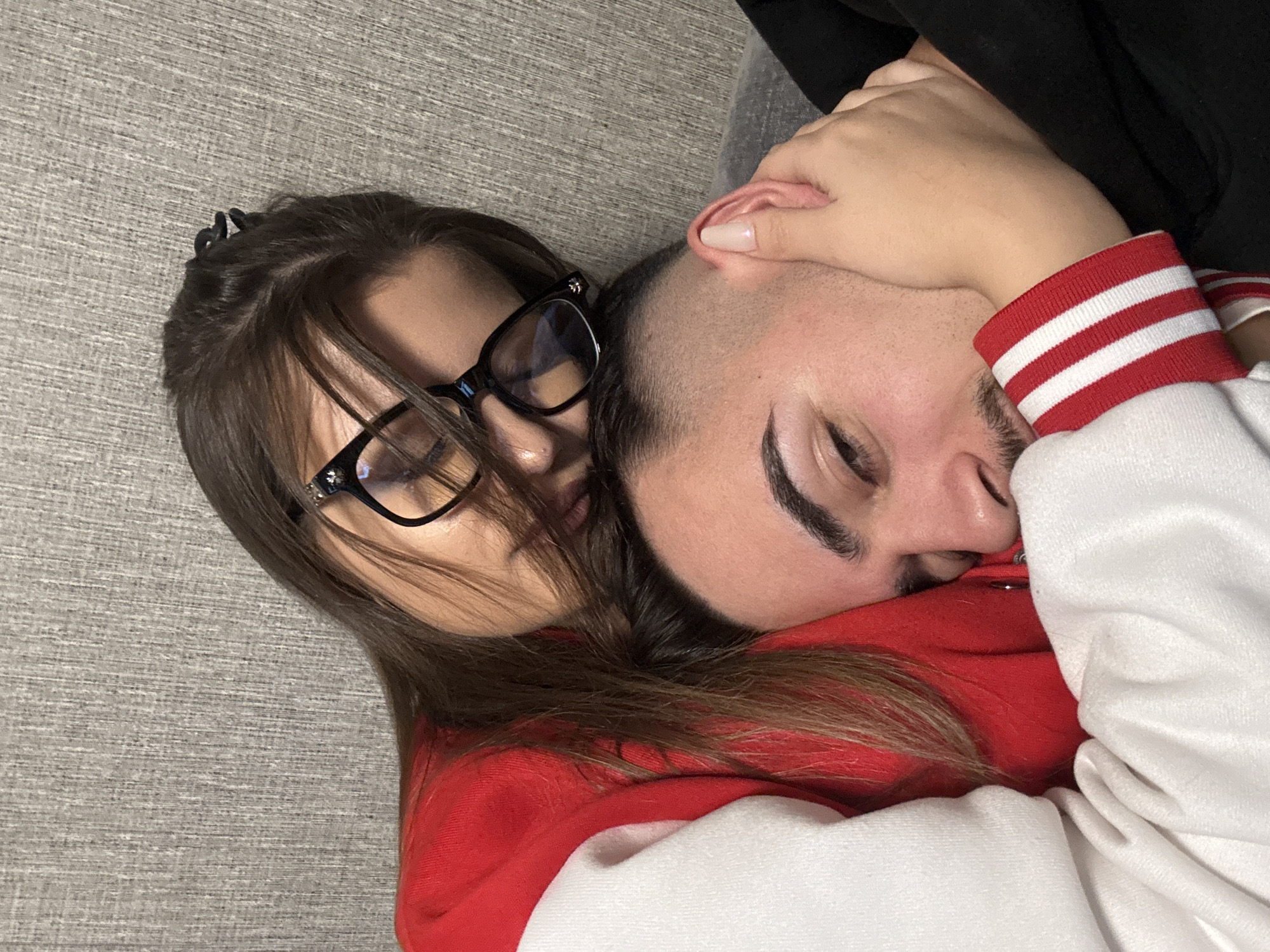 Two individuals are lying down indoors on a gray textured surface, possibly a couch or bed. A young woman with dark hair pulled back with a black clip, wearing black-rimmed glasses, is close to the other individual. The other individual, a male with dark hair cropped short, has his eyes closed and is being embraced. Both are wearing casual clothing; the woman in what appears to be a red sweater, and the male in a white and red striped sweater. The scene is warmly lit. There is no information to tell the location in Sofia, Bulgaria.