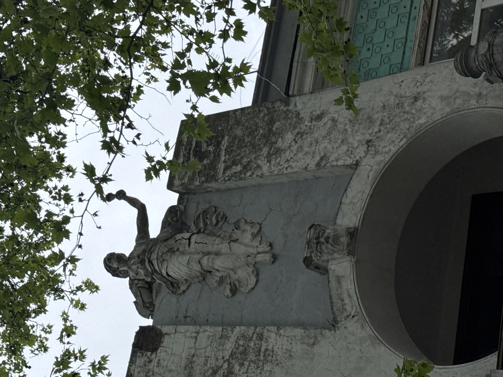 An upward shot captures a section of an ornate building in Yevpatoriya, Ukraine. The building features architectural details such as a semi-circular arch, elaborate moldings, and a sculpted figure holding an object, possibly a fruit or orb. The statue is weathered, showing signs of age and deterioration. The building itself has a textured surface, with cracks and imperfections visible. Overhanging the building are tree branches with dense green leaves, partially obscuring the sky and casting shadows on the facade. Above the arch is a window that features a blue-green decorative grill pattern, adding color to the otherwise muted tones of the building.