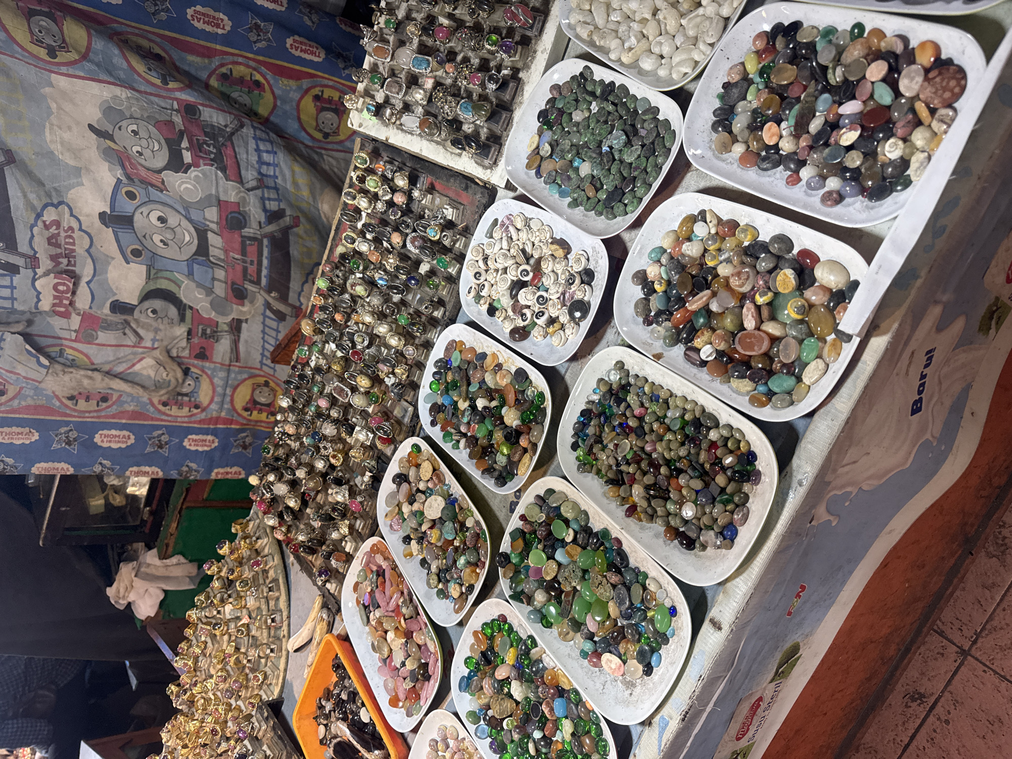 The image depicts a stall at a street market in Denpasar, Indonesia, specializing in gemstones and jewelry. The stall displays a wide array of rings with various colored stones, as well as loose gemstones sorted into white rectangular trays. In the background, a blue tarp decorated with images of Thomas the Tank Engine characters is visible, suggesting a family-friendly market or a stall catering to a diverse customer base. The lighting suggests it is either evening or the stall is under artificial light. The overall impression is one of a vibrant, bustling marketplace.