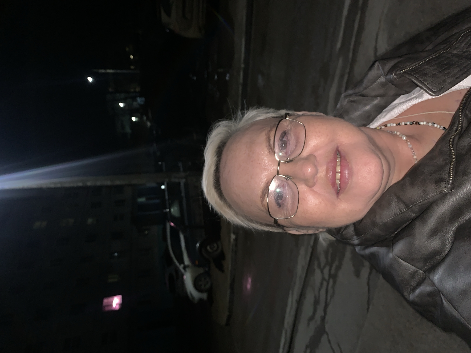 A woman with blonde hair and glasses is facing the camera, wearing a brown leather jacket and necklaces. The background shows an urban environment at night. Streetlights illuminate the scene. A building with lit windows and cars can be seen in the distance. Based on the location tag, this image was taken in Chaikovsky, Russia.