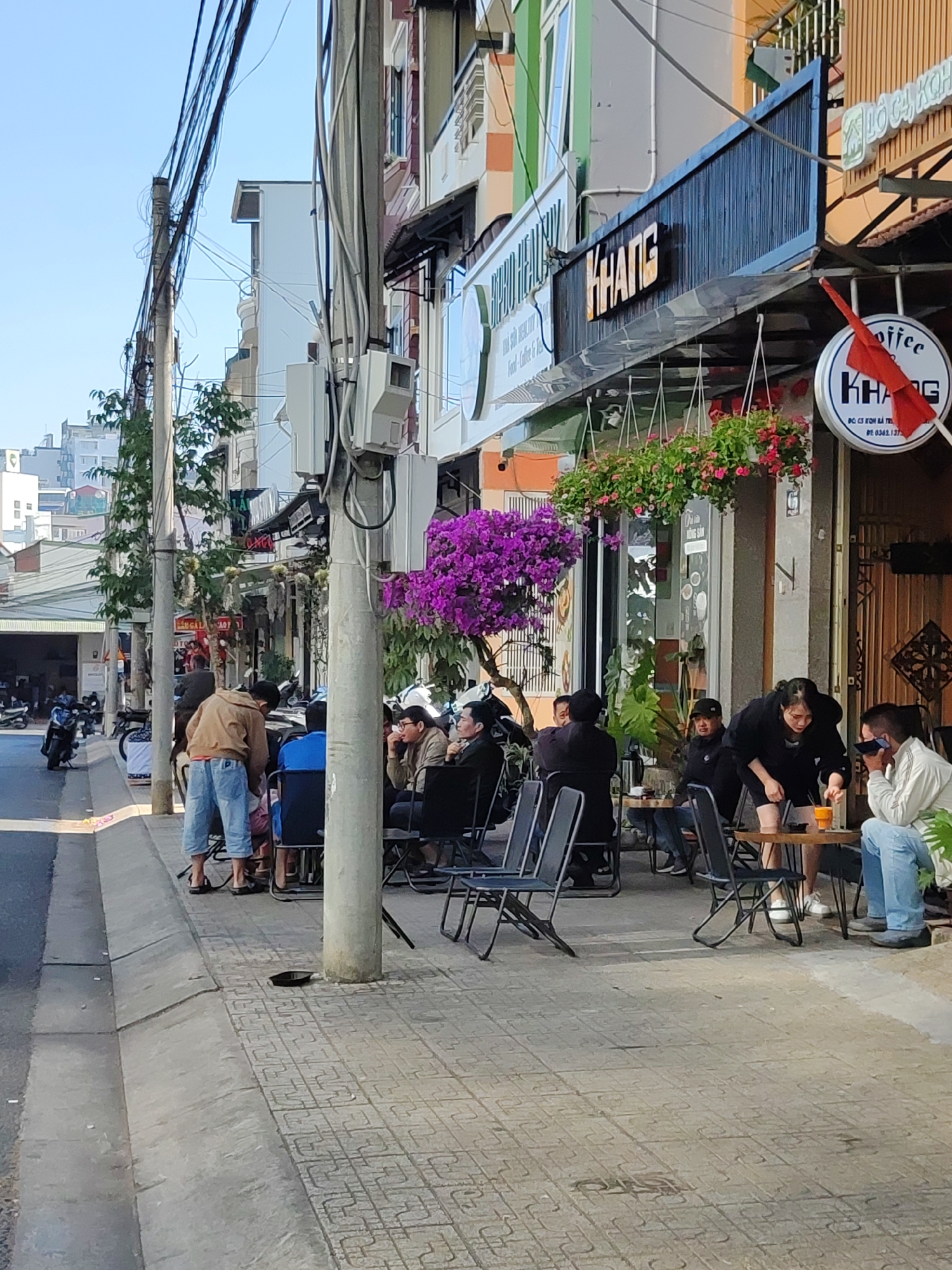 The image depicts a street scene in Da Lat, Vietnam. A group of people is sitting at tables outside a café called "KHANG." Some individuals are looking at their phones, and some are interacting with each other. There are hanging baskets of flowers above the tables and a bougainvillea tree nearby. Motorbikes are parked along the street. Overhead power lines are visible. The sidewalk is paved with patterned tiles.