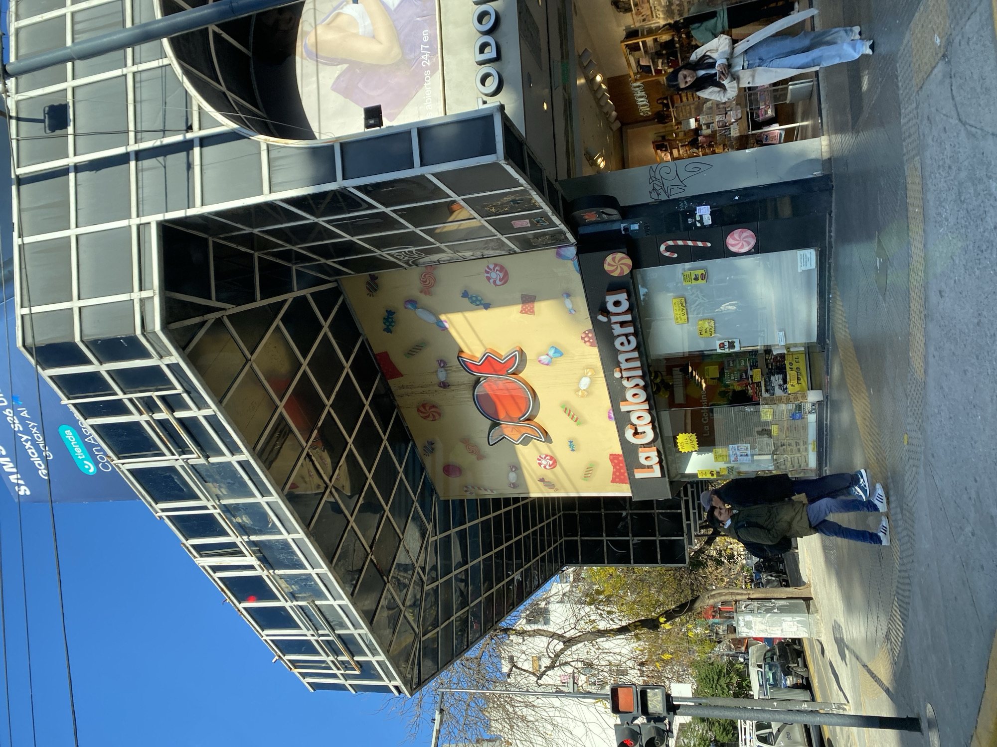 The image captures the exterior of "La Golosineria," a candy store in the city of Buenos Aires, Argentina. The store's facade is decorated with candy motifs and the brand name prominently displayed. A modern building structure with a grid-like pattern surrounds the store. A Samsung advertisement is visible on the left side of the frame, promoting the Galaxy S26 Ultra and Galaxy AI, along with the phrase "Tienda Con Age."
Two individuals wearing jackets, jeans, sneakers and face masks stand on the sidewalk in front of the store. A traffic light is visible in the foreground. The sky is clear and blue. Other commercial establishments are visible in the background.