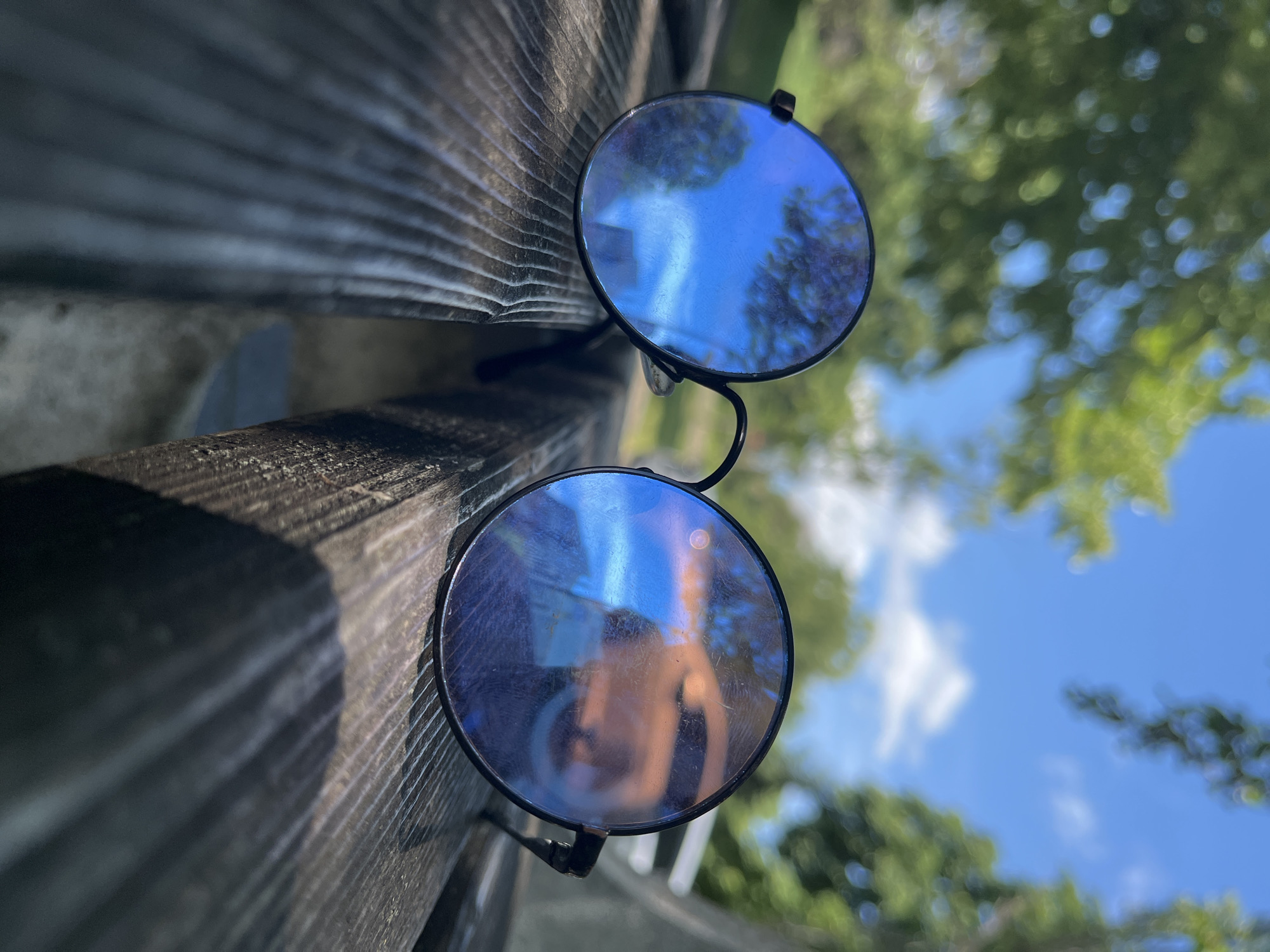 A pair of round, black-framed sunglasses rests on a weathered wooden bench. The lenses are reflective, showing a blurred blue sky with white clouds and green trees. The bench appears to be outdoors, possibly in a park, given the natural backdrop. The lighting suggests it is daytime. There is no indication of the city of Shumen, Bulgaria, within the image.