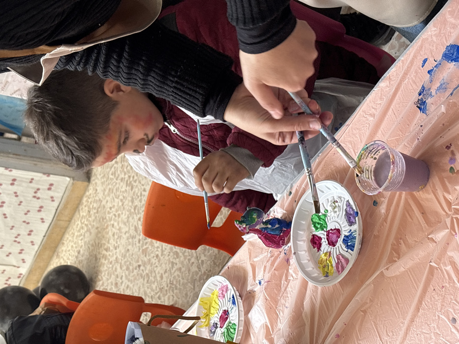 An indoor scene in Kafr Batna, Syria, depicts a creative activity involving two individuals. In the center-left, a young boy with dark hair wears a white protective smock over a maroon zipped jacket. His face is painted with red blush marks on his cheeks and a black mustache above his lip. He holds a paintbrush in his right hand, actively applying paint to a small, multi-colored figurine resembling an animal, positioned on the light pink plastic-covered table in front of him. To the right, another person, largely out of frame, wears a black ribbed long-sleeved garment; their hands are visible, holding two paintbrushes near a clear plastic cup filled with murky purple water, likely for rinsing brushes.
On the table are two white plastic paint palettes, each containing distinct colored paints: yellow, pink, green, white, red, blue, and purple. The foreground palette also holds a paintbrush resting in the green paint. The light pink plastic tablecloth is splattered with various paint colors. In the blurred background, orange plastic chairs are visible, along with a light-colored tiled floor and a wall featuring a patterned design above a white baseboard. Dark, spherical objects are partially visible in the far-left background. The overall scene indicates an organized art activity focused on painting.