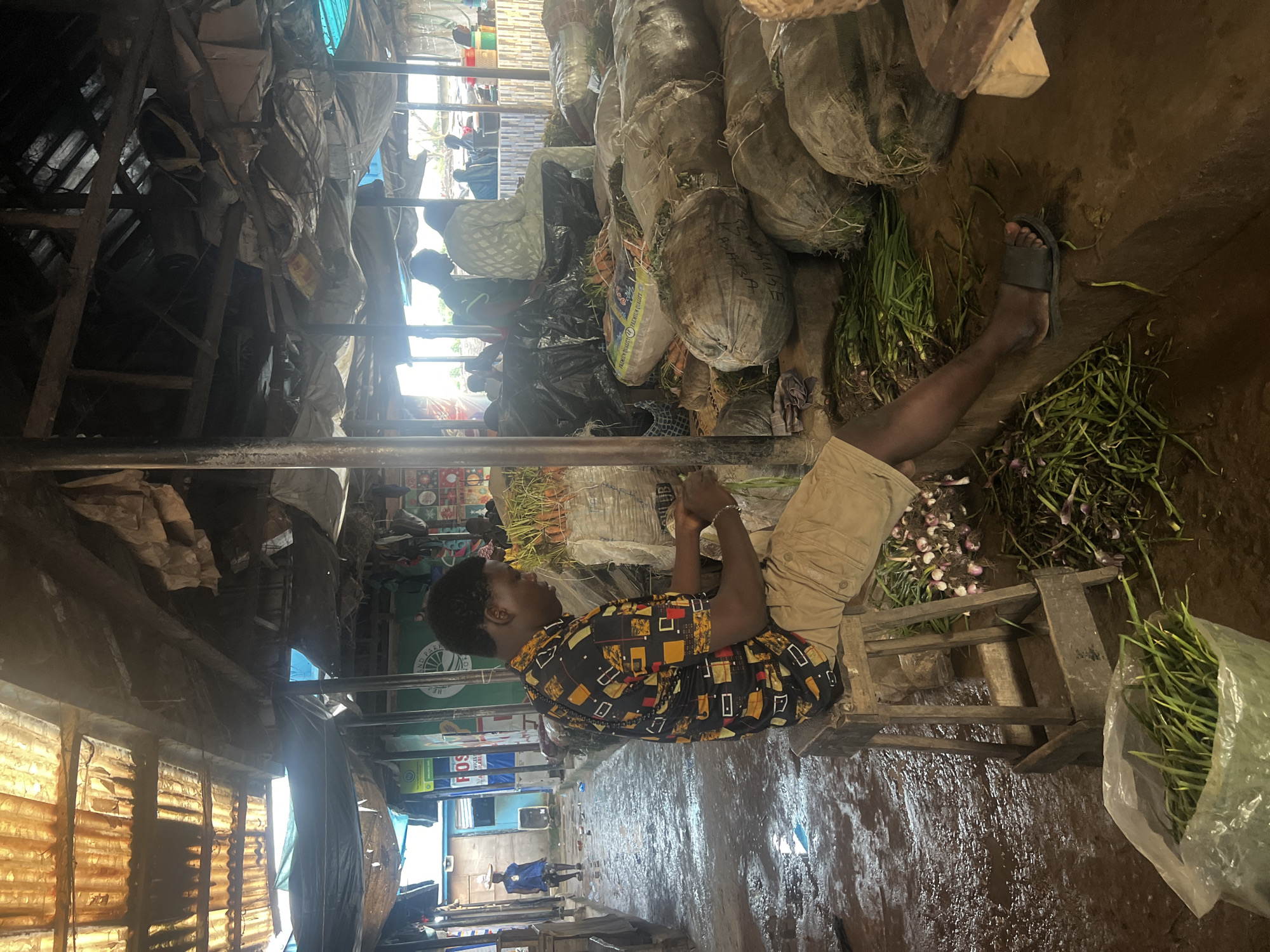 This is a forensic, matter-of-fact description of a scene in Oko Amakom, Nigeria. The image depicts an outdoor market under a makeshift corrugated metal and tarp roof. In the foreground, a young Black man is seated on a wooden stool, wearing a patterned shirt and beige shorts. He is engaged in preparing produce, with bunches of what appear to be onions and green onions scattered around him on the wet, muddy ground. To his right, large sacks, possibly containing produce like potatoes or yams, are stacked. In the background, other individuals are visible, some seated and others standing amidst stalls and more sacks. A woman in traditional attire walks in the distance, carrying a load on her head. The environment is characterized by natural light filtering through the worn roofing, casting shadows and highlighting the dampness of the ground. The overall impression is one of a busy, functional marketplace.