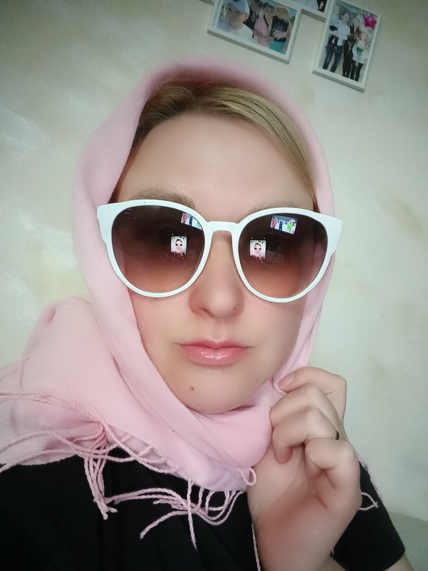 A woman wearing a light pink headscarf and white-rimmed sunglasses is posing for a selfie in Chaikovsky, Russia. The frames on the wall in the background possibly contain other pictures of the woman and possibly her friends. The reflection in the sunglasses appears to be of the woman holding a cell phone, taking a picture of herself. The woman is wearing a black shirt. The overall lighting of the photo appears to be even and diffused. The walls are a neutral color.