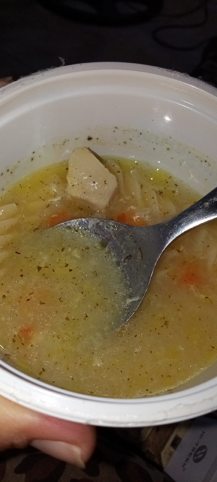 A close-up shot reveals a bowl of soup. A metal spoon rests in the soup, partially submerged. The soup appears to contain pasta, diced carrots, potatoes, and green herbs. The bowl is white and has a slightly rough rim. The overall lighting is dim, casting shadows in the background. A hand is visible along the lower left portion of the frame, and what appears to be some type of electronic device with writing is visible in the bottom right corner of the shot.