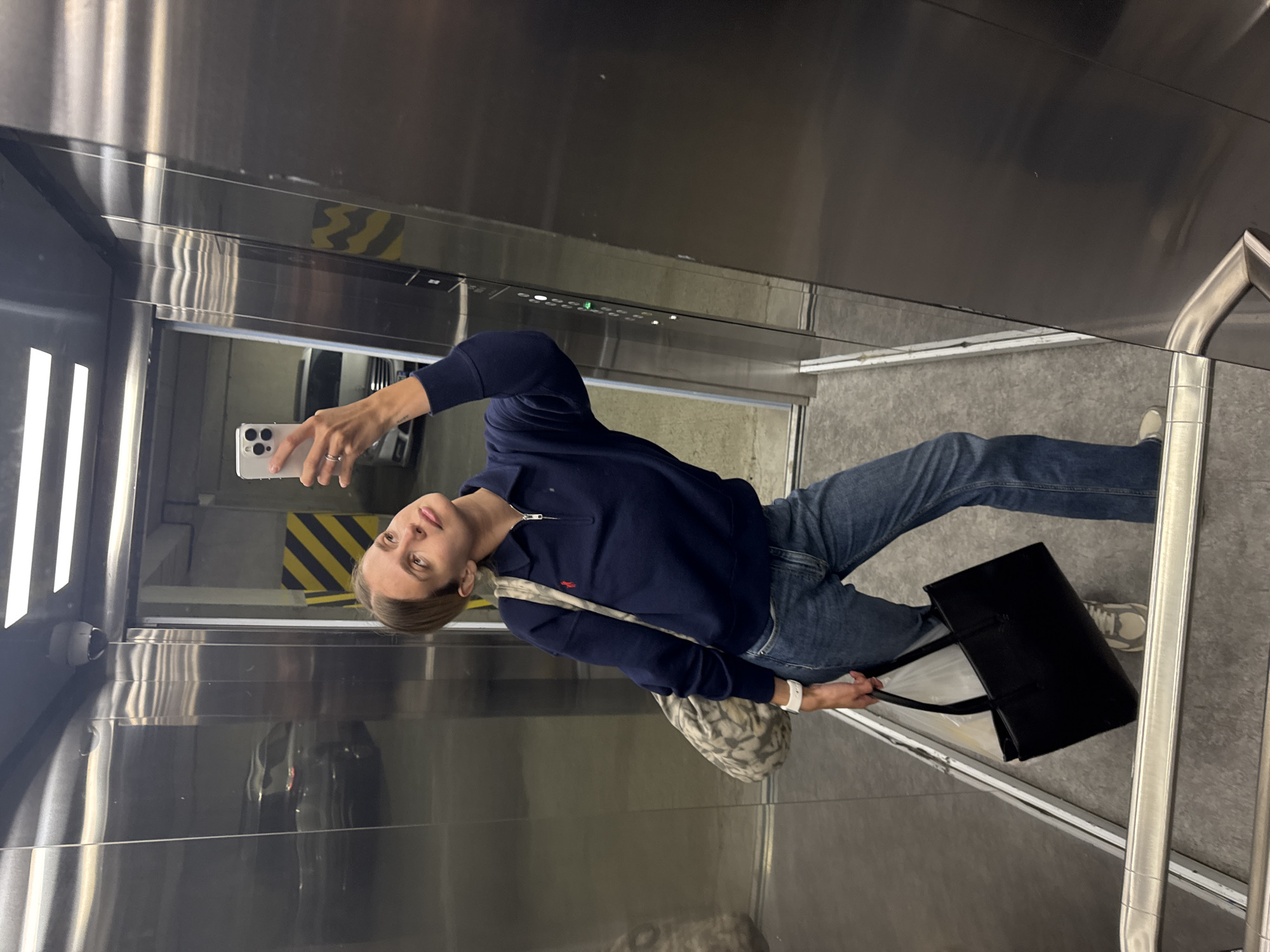A person is taking a selfie in a metal elevator. The person is wearing a dark blue pullover, blue jeans, and white sneakers. They are holding a white smartphone in their left hand and a black tote bag in their right hand. They also have a patterned shoulder bag. The elevator walls are made of brushed metal and have a row of buttons. The floor is gray. There is a yellow and black hazard stripe visible on the wall behind the person. A metal handrail is visible on the right side of the elevator.