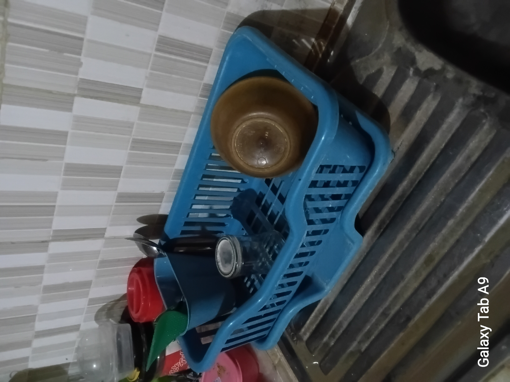 This image depicts a blue plastic dish rack situated on a gray sink in what appears to be a kitchen environment. The dish rack contains a clear glass, a brown bowl, and a green spatula. Behind the dish rack, a tiled wall with alternating light and dark gray stripes is visible. There are no people or animals present in the image. The lighting suggests an indoor setting, likely during daylight hours or with artificial lighting. The device information at the bottom of the image indicates it was captured on a "Galaxy Tab A9", which is a model of tablet computer. It is not possible to determine the specific location within the city of Trenggalek, Indonesia from the provided image.