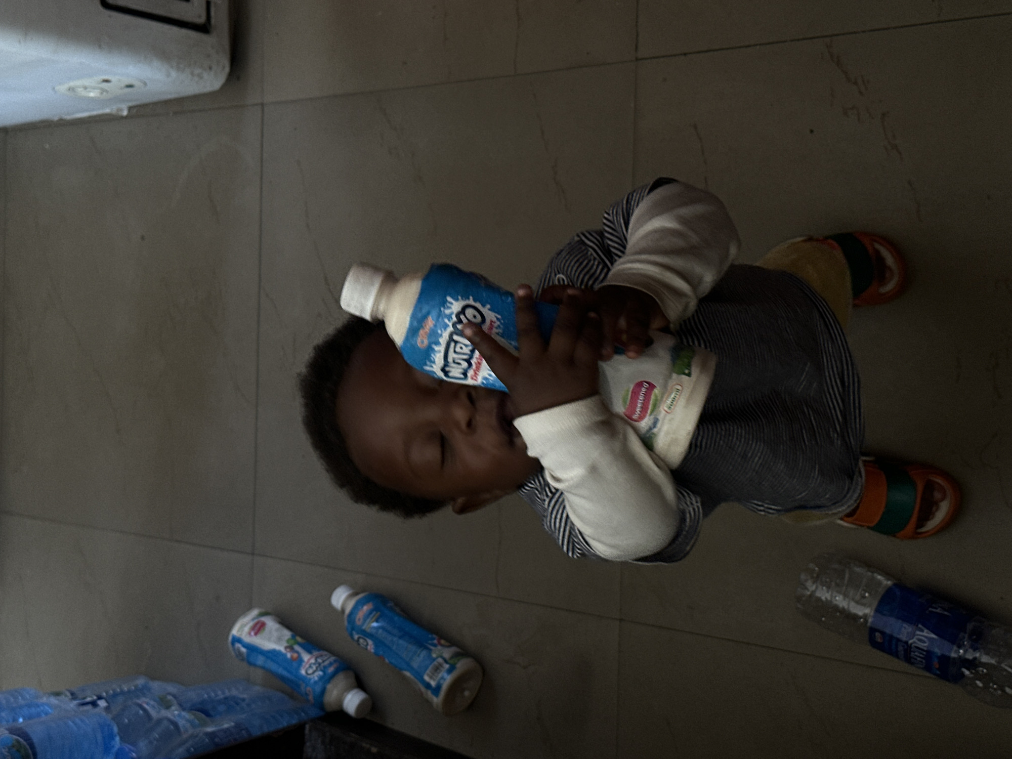 In this vertically oriented, indoor photograph taken in Gura Top, Nigeria, a young Black child is the central subject. The child is dressed in a light-colored, long-sleeved shirt with thin blue stripes. The child is actively drinking from a blue and white plastic bottle labeled "Nutri-O Drink" in white lettering with a blue outline and a white cap. The child's eyes are closed, and their head is tilted back, suggesting absorption in the act of drinking.
The child is positioned on a tiled floor, which appears to be a light grey or beige with a subtle marble pattern. To the child's left and slightly behind them, several other bottles are visible. These include two more blue and white "Nutri-O Drink" bottles and a clear plastic bottle of water labeled "Aquafina." A portion of a white appliance with visible electrical outlets is also in the upper left corner of the frame. The overall lighting suggests an indoor setting, possibly during the day, but without strong direct sunlight. The mood conveyed is one of quiet, focused consumption by the child.