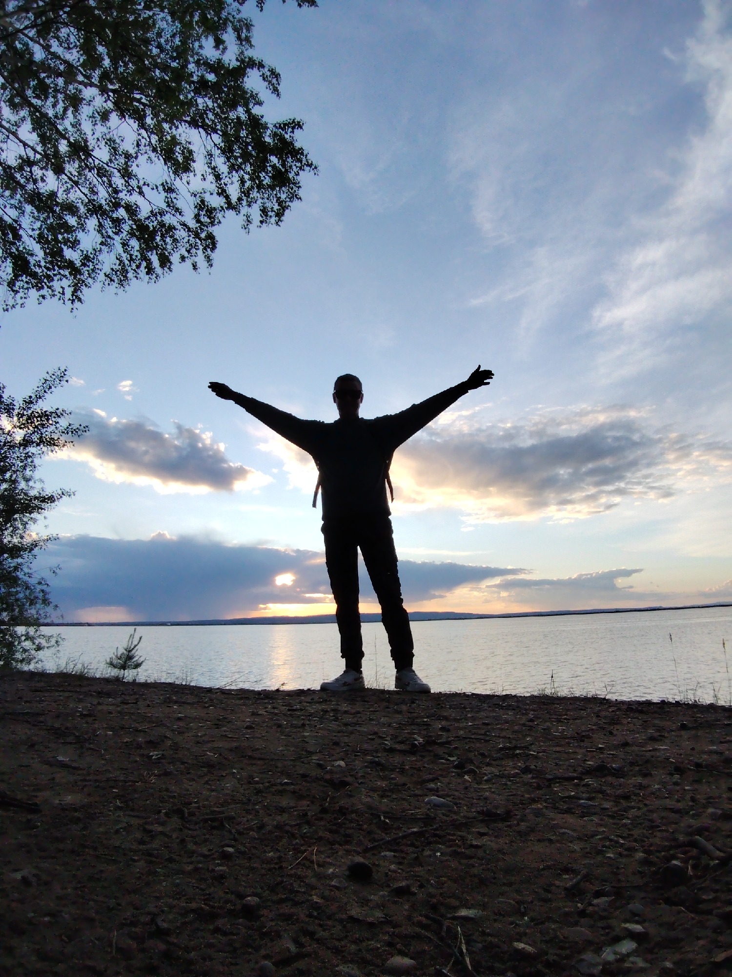 A silhouette of a person stands with arms outstretched on a dirt surface, overlooking a body of water. The sky displays a sunset or sunrise, with clouds scattered across. To the left, a tree branch with leaves partially frames the scene. The location is Chaikovsky, Russia.