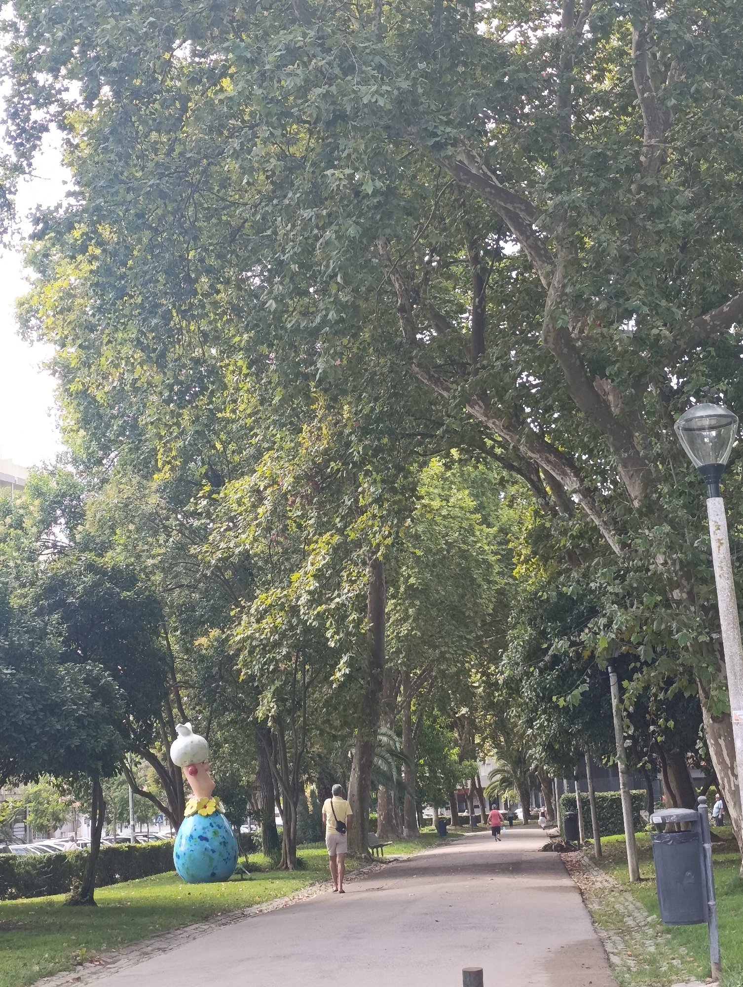 The image depicts a tree-lined park pathway in Setúbal, Portugal. On the left side of the path, a whimsical statue of a figure with a large, blue, egg-shaped body and a white, bulbous head is visible. The statue is adorned with yellow flowers around its neck. A person wearing a yellow shirt and light-colored shorts is walking away from the statue on the path. Further down the path, other individuals can be seen walking, and on the right side, a lamppost and trash receptacles are present. The pathway itself is paved and curves slightly, bordered by grassy areas and abundant trees with lush green foliage. In the background, a street with parked cars is discernible.
