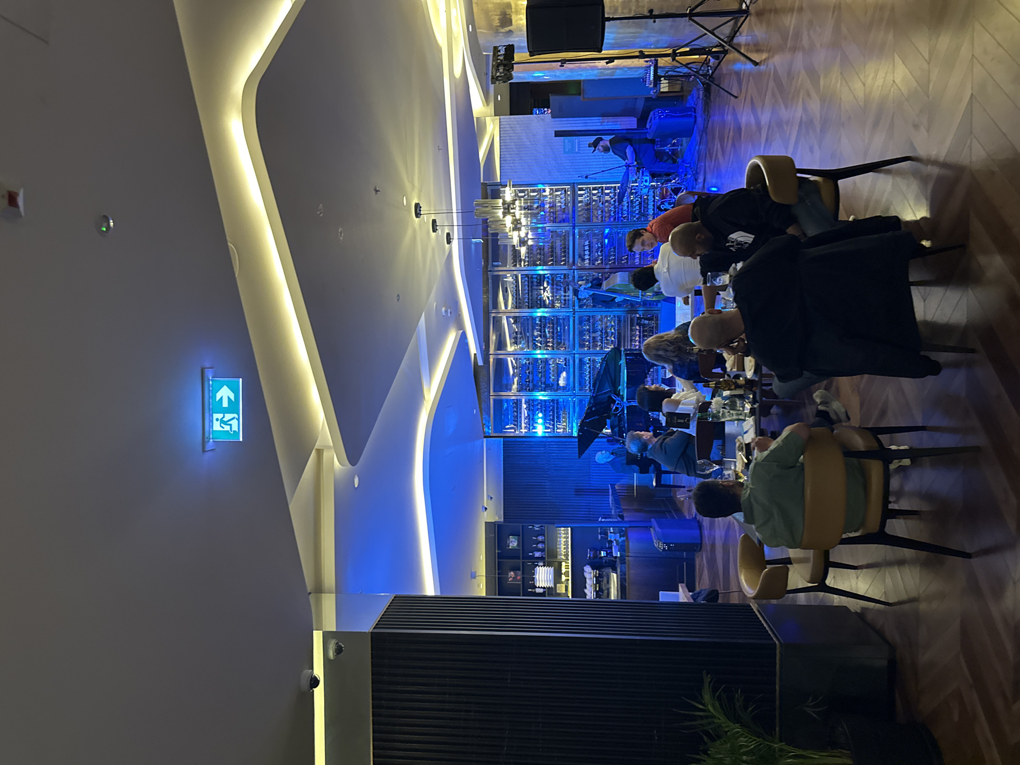 The image depicts the interior of a bar or restaurant in Batumi, Georgia. The setting features contemporary design, with a focus on blue and gold lighting. A live band is performing in the background near a well-stocked bar with numerous bottles. Several patrons are seated at tables, engaged in conversation. The furniture is modern, and the floor has a herringbone pattern. An exit sign is visible on the wall near the entrance. The scene captures a social gathering in an upscale establishment.