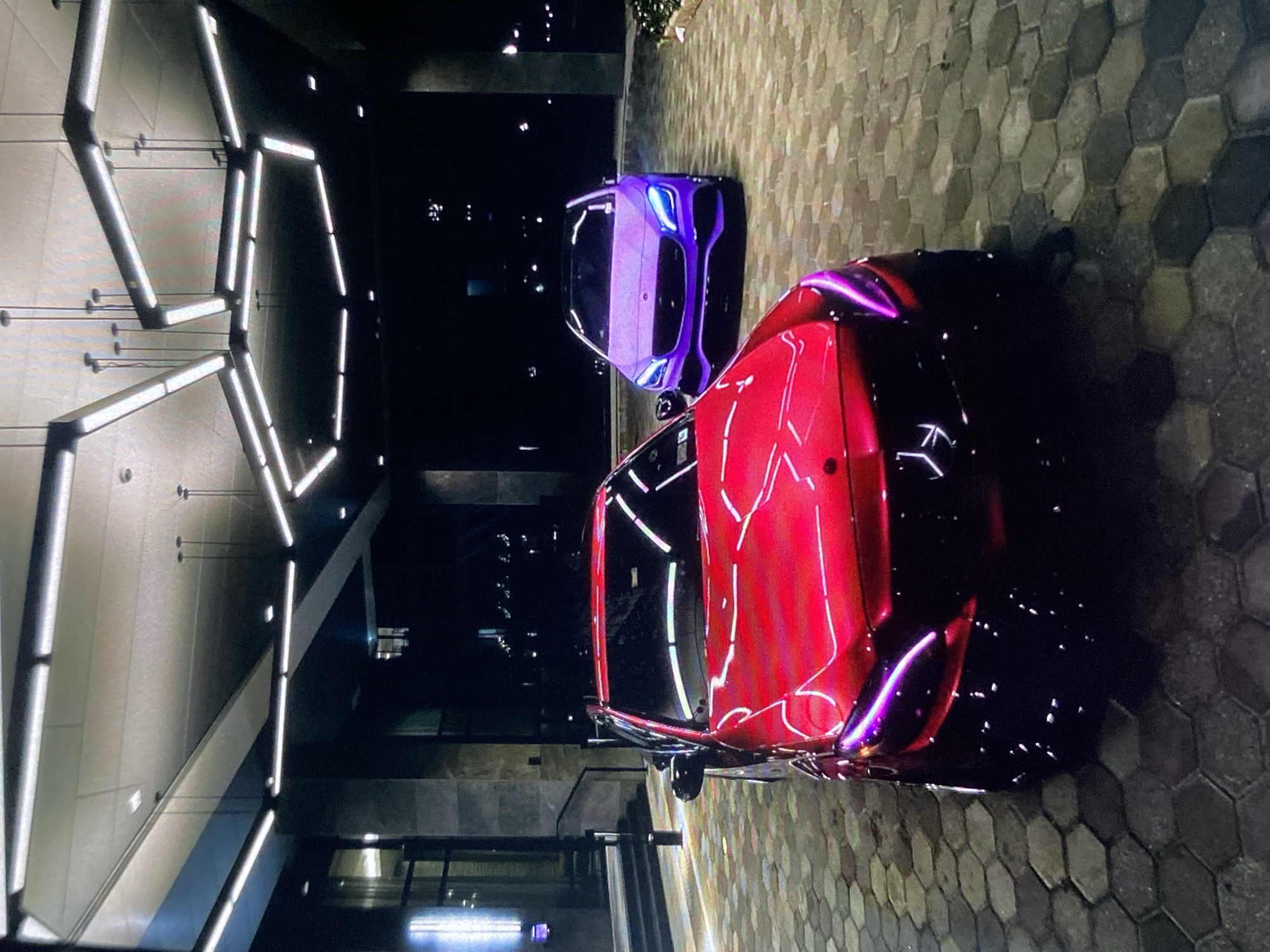 The image depicts two cars parked on a patterned stone surface. The foreground car is red, reflecting light, with purple accents. Behind it is a purple car. The location appears to be an urban setting at night. In the background, there are city lights and what appears to be a modern building with geometric lighting fixtures. There are no people visible. There is no information indicating that this is in Lhokseumawe, Indonesia.