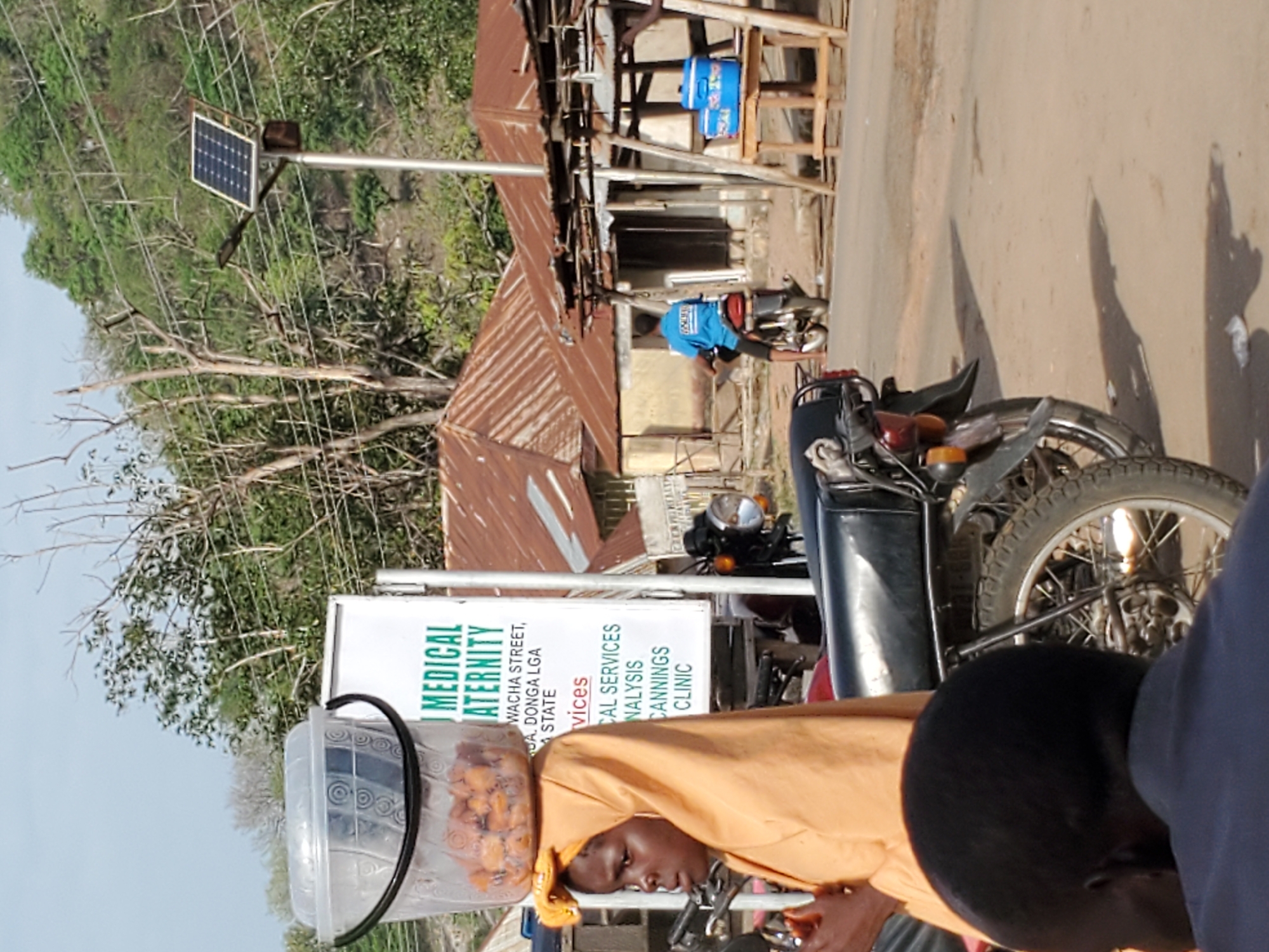 This image captures a vibrant street scene in Mararaba, Nigeria, under bright, sunny daylight, evidenced by the strong shadows cast on the ground. In the foreground, a person, likely a woman, wearing an orange headscarf and top, carries a large, clear plastic container filled with light brown, spherical snacks or edibles balanced on her head, indicative of a street vendor. Part of another person's head and shoulder are visible in the extreme right foreground.
In the mid-ground, a black motorcycle is parked, and another individual in a blue shirt is seen near a different motorcycle or structure. The background features buildings with characteristic rusty corrugated iron roofs and wooden support structures, common in the region. A tall pole with a solar panel and an attached street light stands prominently, suggesting the use of solar energy for public lighting. Several blue plastic containers are visible on a wooden stand near one of the buildings.
A white signboard is clearly visible, displaying text that reads: "MEDICAL MATERNITY", followed by an address "WACHA STREET, DA. DONGA LGA STATE". Further text lists "services" such as "CAL SERVICES", "NALYSIS", "CANNINGS", and "CLINIC", indicating a medical facility. The surrounding environment includes green foliage and trees, adding to the semi-urban or rural feel of the location. The image depicts a slice of daily life and commerce on a local street.