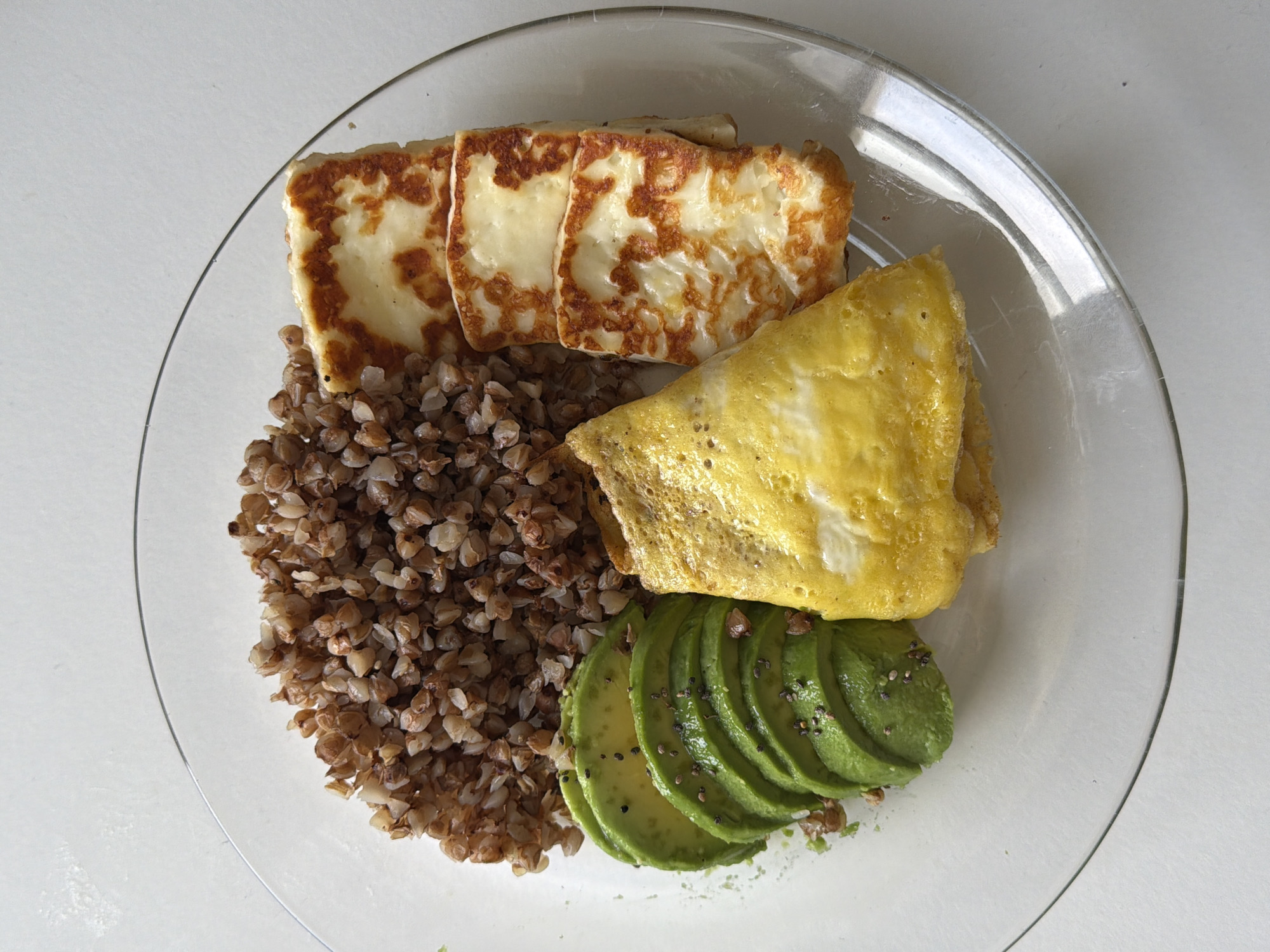 A clear glass plate sits on a white surface, containing a portion of cooked buckwheat, several slices of avocado sprinkled with black seeds, a folded omelet, and several slices of grilled halloumi cheese. There are no people present. The setting does not allow for identification of Tel Aviv.