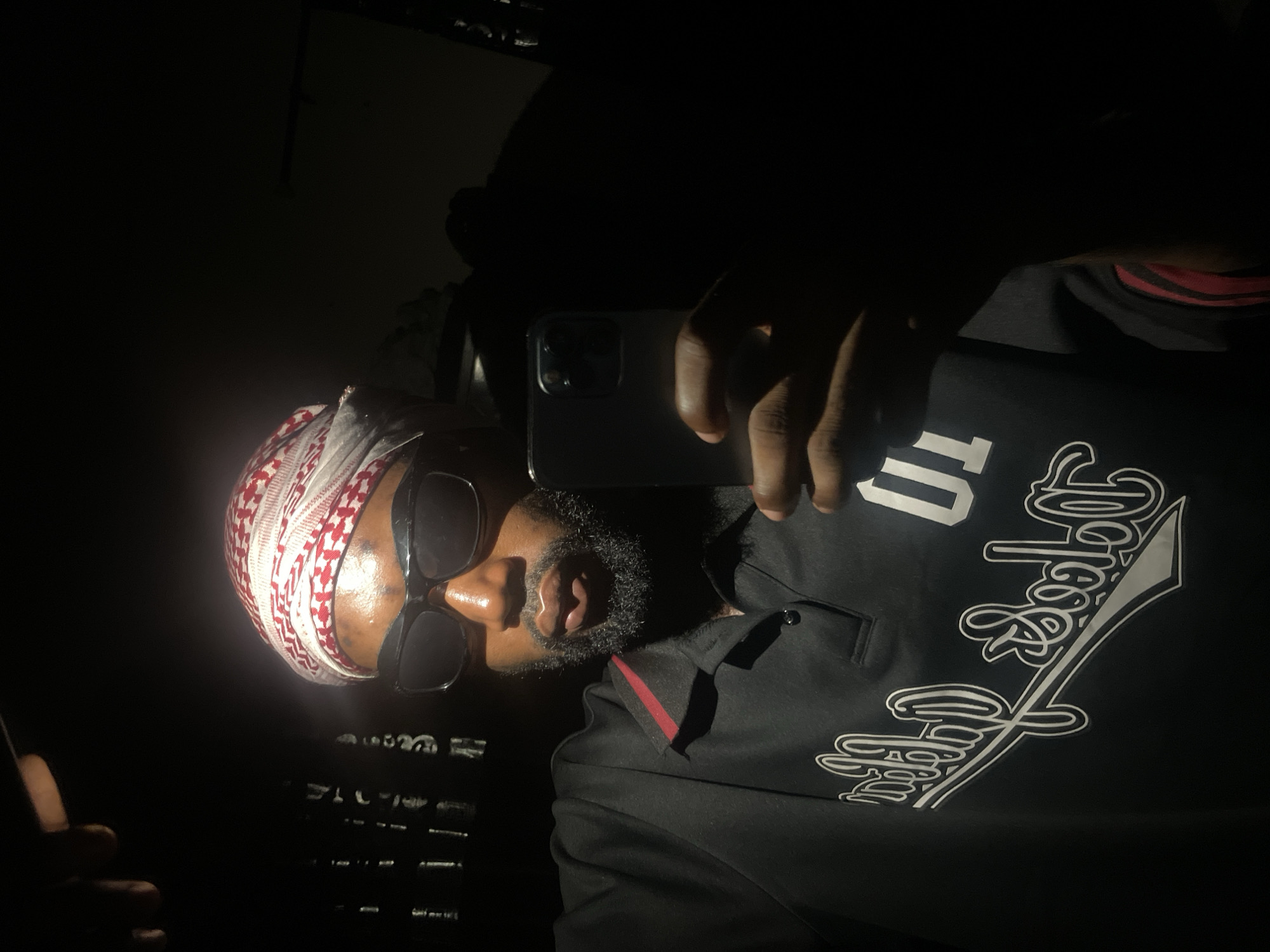 A medium, dark, indoor selfie shows a Black man wearing a white and red checkered headdress, sunglasses, and a black jacket with white decorative lettering and number. He is holding a cell phone and looking at the camera. The background is very dark with light shining directly on the man's face and part of his body. The location, Lokoja, Nigeria, cannot be determined from the image.