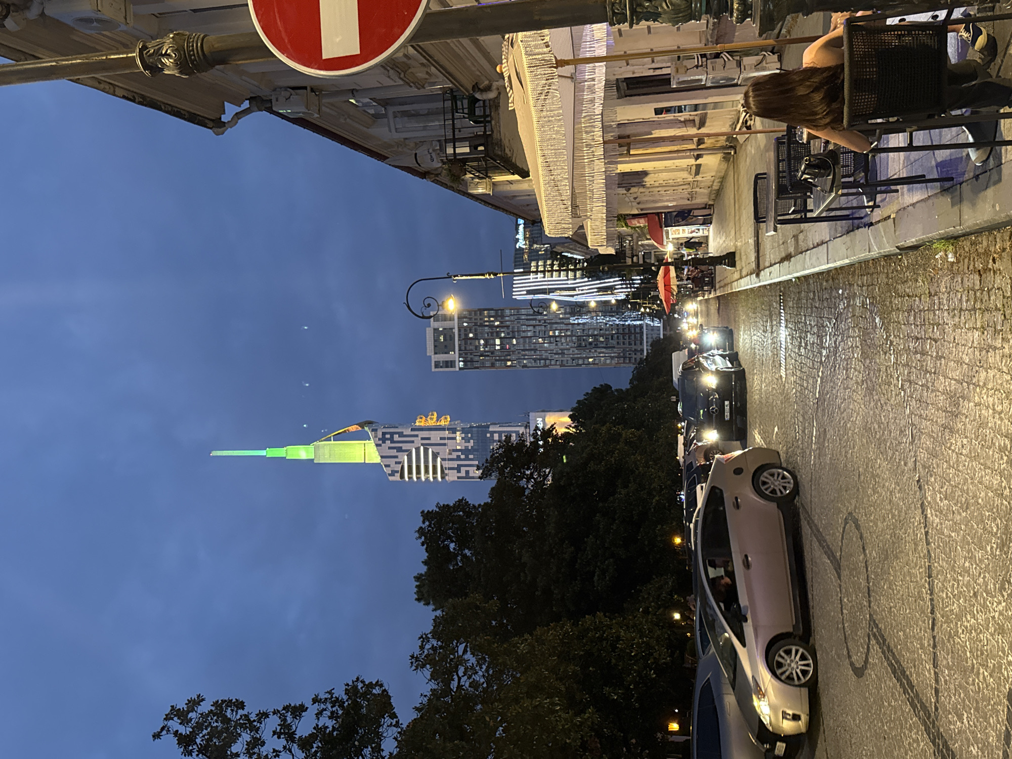This image captures a street scene in Batumi, Georgia, at dusk. The view is from a street level perspective looking down a cobblestone road lined with buildings and trees. In the distance, a tall, modern skyscraper with green and white lighting dominates the skyline, its pointed spire reaching into the darkening sky. Another, shorter skyscraper with many illuminated windows stands to its left, and further down the street, a building with a red roof is visible.
On the right side of the street, an older building with ornate architectural details features a balcony and a white awning extending over an outdoor seating area. Several tables and chairs are set up on the sidewalk, with a woman seated at one, facing away from the camera. A circular red and white sign, likely a traffic sign, is prominent in the upper right corner of the image.
The street itself is paved with cobblestones, and several cars are present, including a silver hybrid vehicle in the foreground and a black car with illuminated headlights further down the road. Trees with dark green foliage are situated along the left side of the street, partially obscuring the view of some buildings. The overall atmosphere is that of a city street transitioning from day to night, with artificial lights beginning to illuminate the urban landscape.