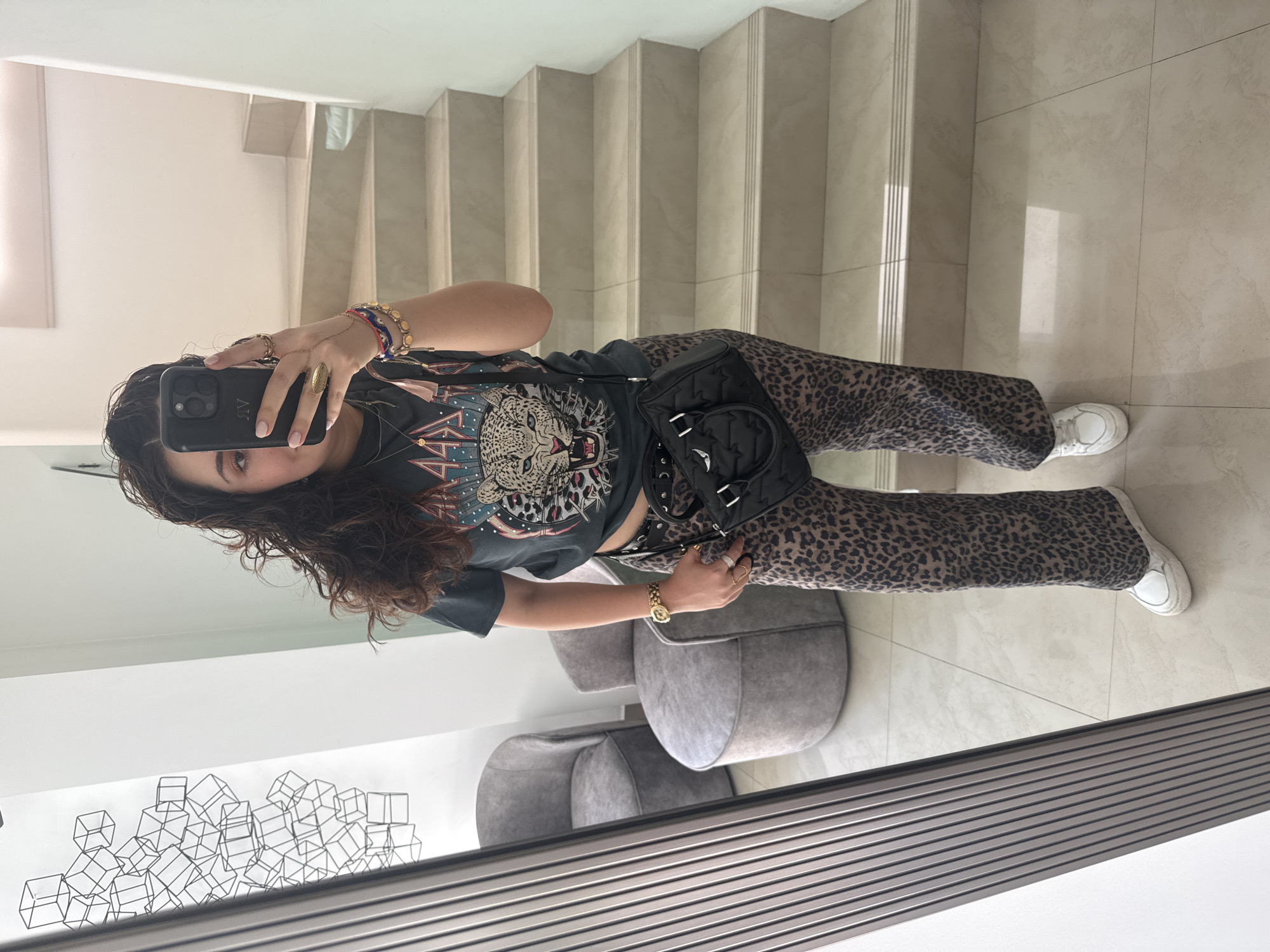 A woman is taking a mirror selfie indoors. She is wearing a graphic t-shirt with a leopard print design, leopard print pants, and white sneakers. She has a black purse slung over her shoulder and multiple bracelets on her wrist. Her hair is dark and curly. The background includes a staircase with light-colored treads, a tiled floor, two gray rounded chairs, and a geometric art piece on the wall. The image appears to be taken in a home setting. There is no way to confirm this is Santa Clara Ocoyucan, Mexico.