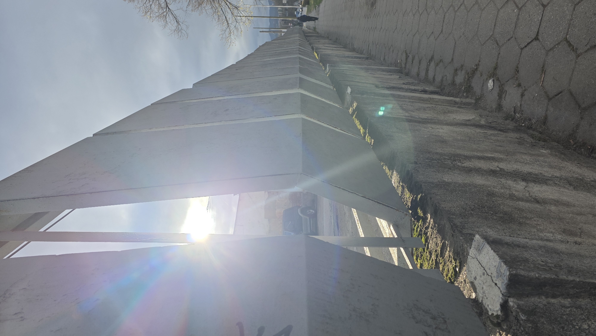 The image shows a perspective view along a concrete wall structure in Blagoevgrad, Bulgaria. The structure consists of repeating triangular-topped segments. A sidewalk runs alongside the wall, constructed of hexagonal pavers. There is a person further down the walkway. The sun is positioned behind the wall, creating lens flare and bright spots. Through gaps in the structure, a car is partially visible. Bare tree branches are visible against a sky with light clouds at the top of the image.