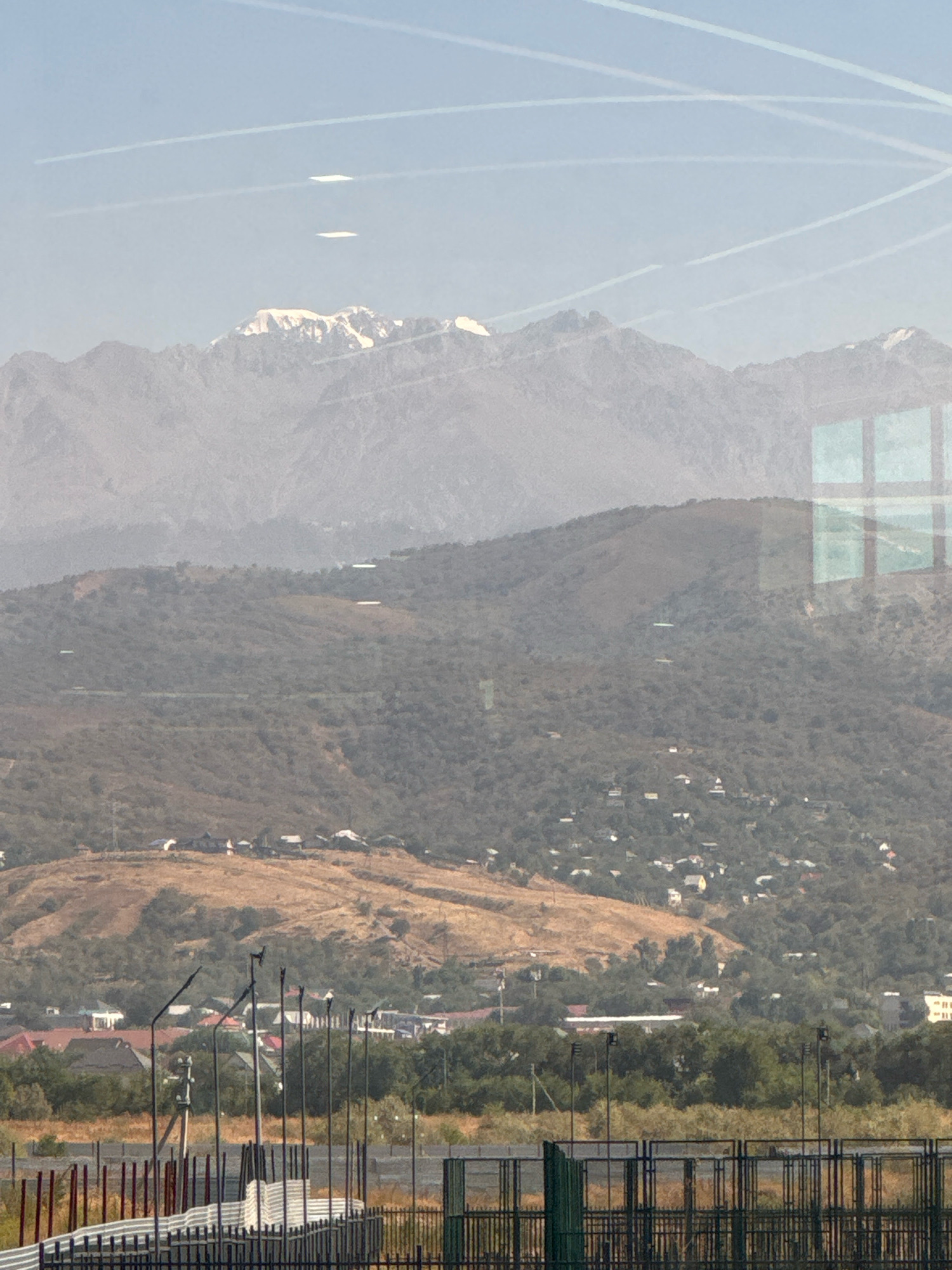 This is a long-range, slightly hazy photograph of a mountainous landscape with a settlement in the foreground. In the far distance, snow-capped mountains are visible against a pale blue sky. There are multiple white contrails crisscrossing the sky, indicating airplane activity.
In the mid-ground, the slopes of the mountains are covered in dense green vegetation and trees. Scattered throughout this area are small white structures, likely houses, forming a hillside village. A distinct ridge of dry, brown grass separates the upper slopes from the lower settlement.
The foreground of the image features a network of dark metal fences and poles, suggesting a constructed area like a sports field or industrial site. Several lampposts are also visible. The overall impression is of a semi-rural or exurban area adjacent to a mountainous region. The presence of contrails suggests this is an area with air traffic, common for a city like Almaty, Kazakhstan, which is known for its proximity to the Tien Shan mountains. The faint reflection on the left side of the image indicates the photograph was taken through a window.