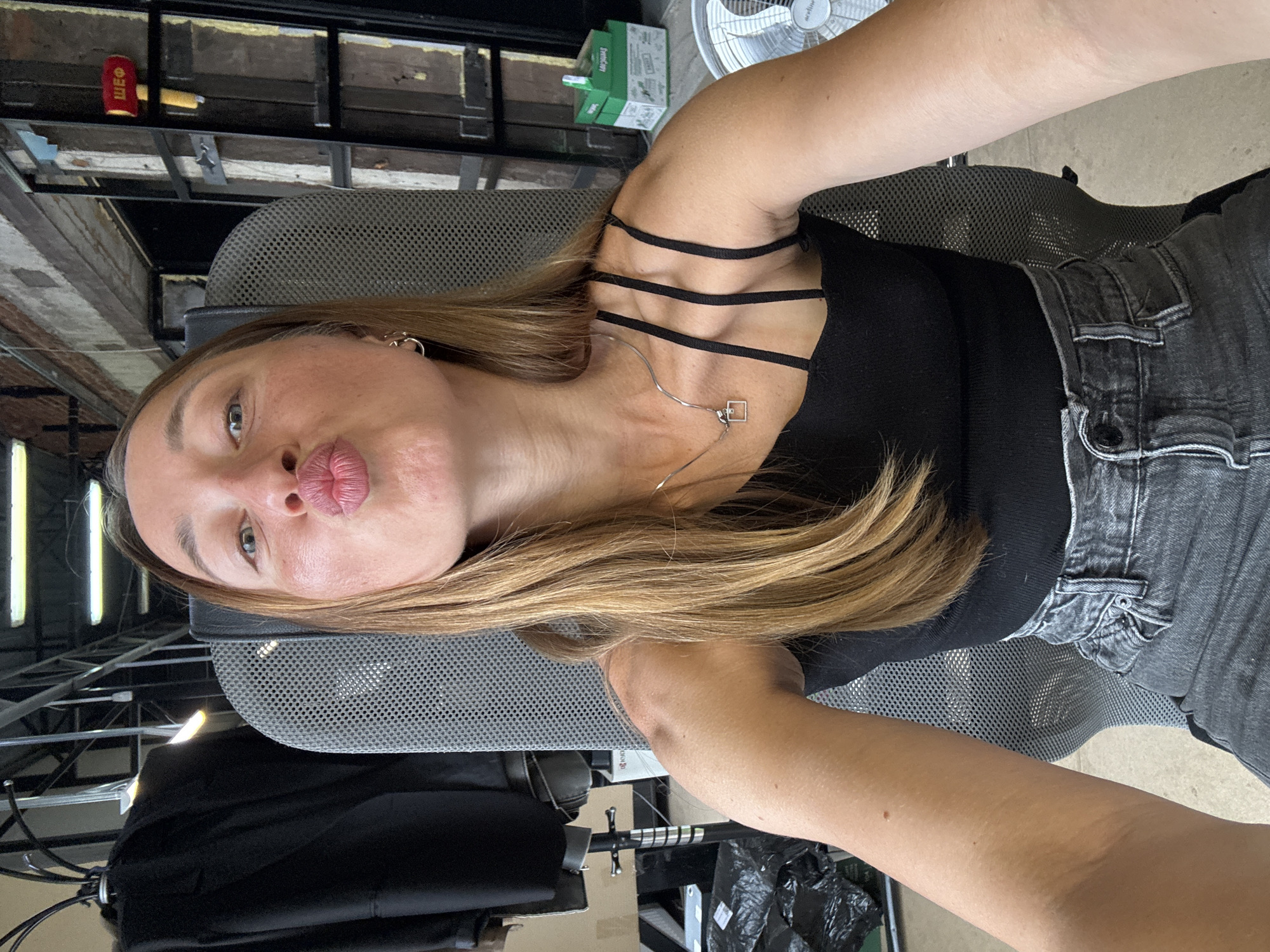 A woman with light brown hair puckers her lips in a selfie. She wears a black top with straps and gray jeans. She sits in a gray mesh office chair. The background includes a cluttered industrial or office space. Items such as a fan, boxes, and exposed ceiling infrastructure are visible. There is no information available from the image to verify it is located in the city of Chaikovsky, Russia.