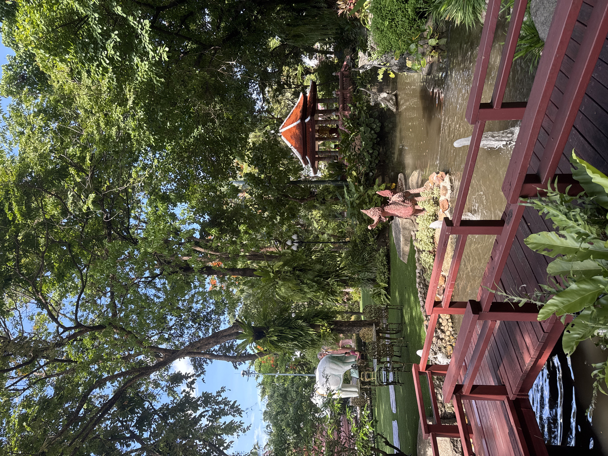 The image depicts a serene outdoor scene, likely a garden or park setting in Samrong Nuea Subdistrict Municipality, Thailand. A pond or small lake is visible with a decorative gazebo or pavilion situated on its bank, surrounded by lush greenery and trees. A wooden bridge with red railings crosses part of the water feature. Manicured lawns and ornamental sculptures are visible. The sky is partially visible through the trees, indicating a bright day.