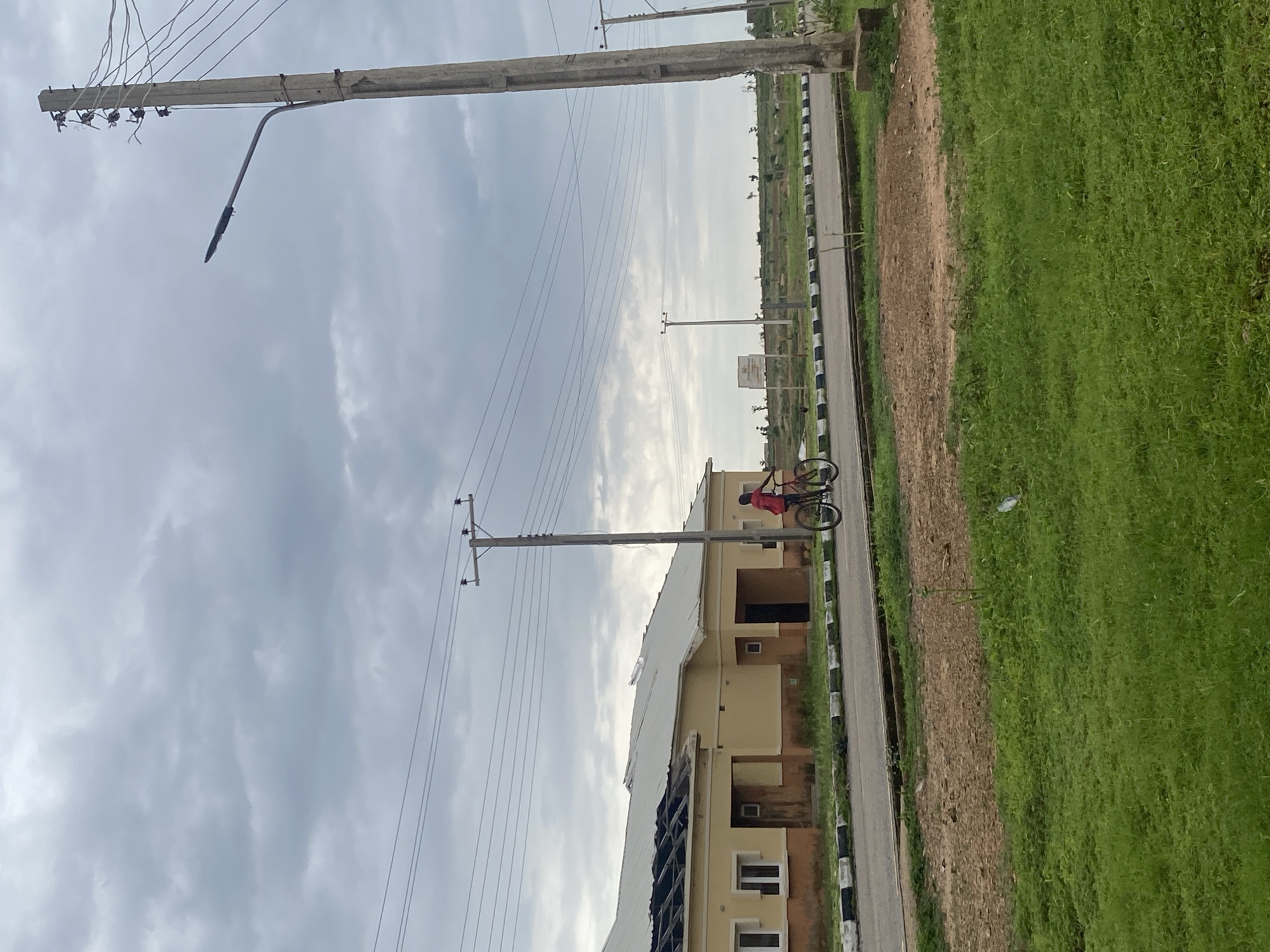 This is a vertically oriented, eye-level photograph taken outdoors in Bulamare, Nigeria. The scene depicts a residential area under a cloudy sky. The foreground features a road with black and white curbing, bordered by a strip of brown dirt and then a lush green grassy field.
The main subjects are two people on a bicycle. One person is actively riding the bicycle, while the other appears to be sitting on the back. They are both dressed in casual clothing. The rider is wearing a red top.
In the mid-ground, there is a light-colored building with multiple windows. The roof of the building appears to be damaged or under construction, with some charred areas visible. Several utility poles with electrical wires are prominent throughout the image, suggesting the presence of infrastructure.
The sky is overcast with a mix of gray clouds, indicating it might be a cloudy or possibly rainy day. The lighting is diffused, suggesting it is either early morning or late afternoon. The overall setting appears to be a developing or partially constructed residential area, possibly a housing estate. There is no visible text in the image.